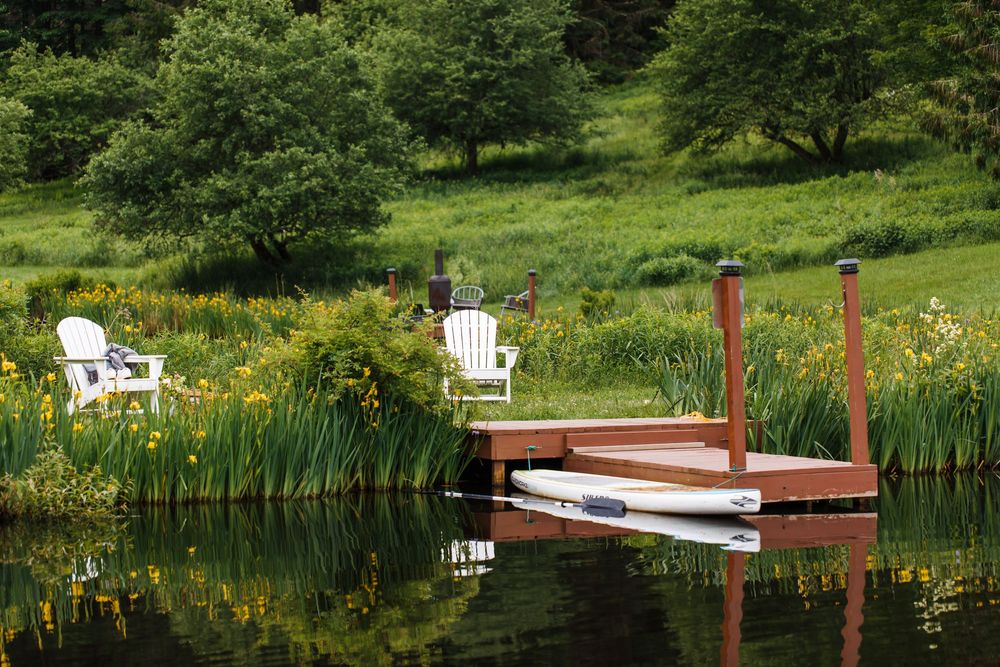 Serene lakeside dock with white Adirondack chairs surrounded by lush greenery and yellow wildflowers reflecting on calm water.