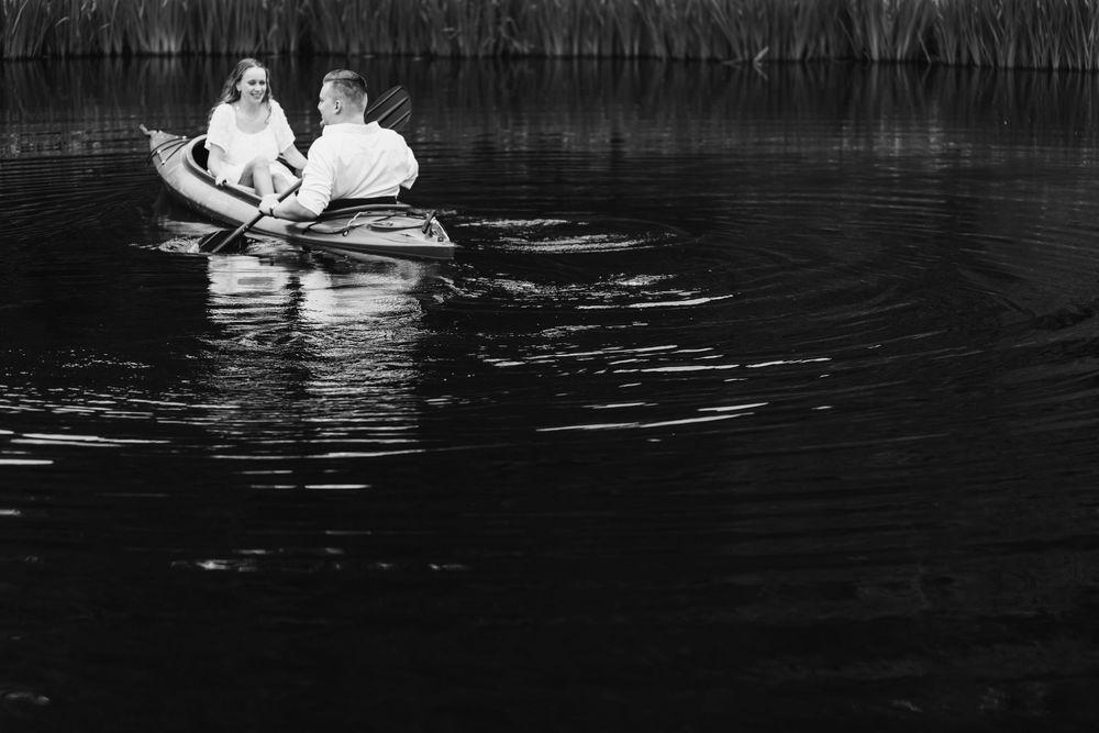 Black and white photo of a couple rowing a small boat on a calm lake surrounded by reeds.