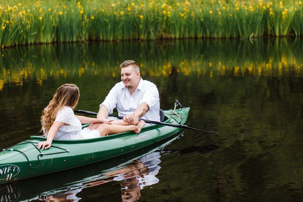 Couple holding hands in a green kayak on a calm lake surrounded by lush yellow wildflowers.