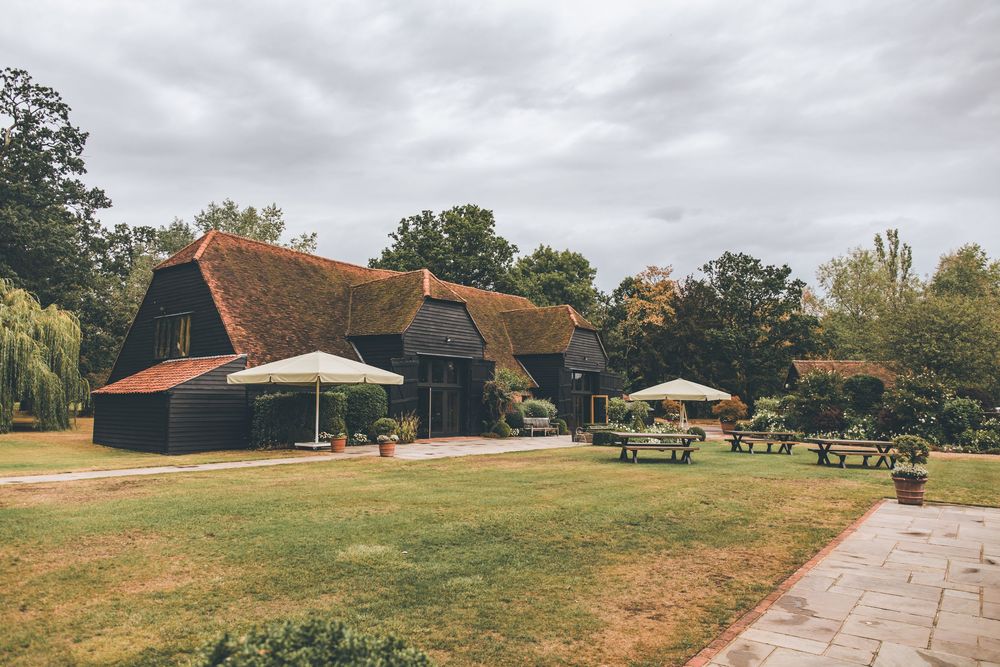 Rustic barn venue with terracotta roof, black timber walls, outdoor seating, and lush green lawns surrounded by trees.