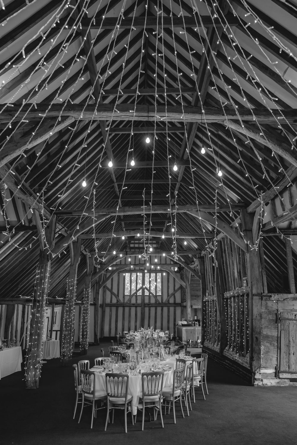 Black and white photo of an elegant barn wedding venue with fairy lights, exposed timber beams, and round dining tables set for a reception.
