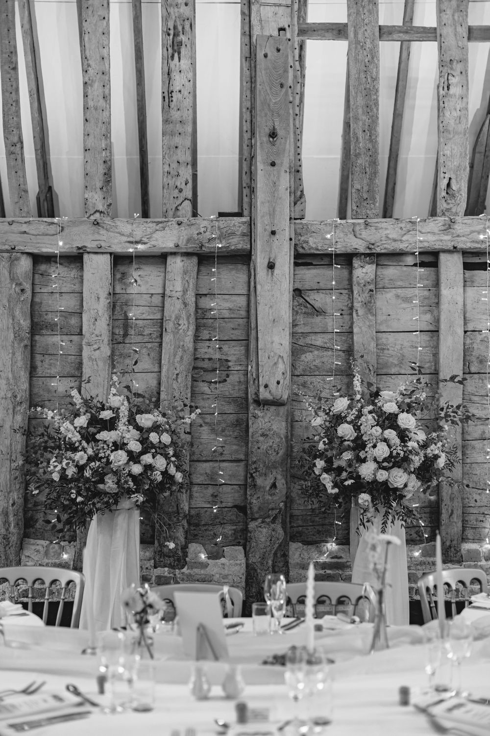Black and white photo of rustic barn wedding reception with large floral centerpieces and fairy lights on wooden beams.