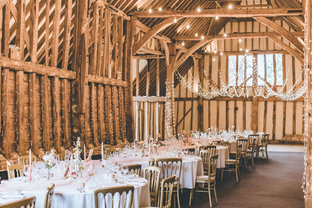 Rustic barn wedding venue with string lights, white round tables, gold chairs, and elegant floral decorations.