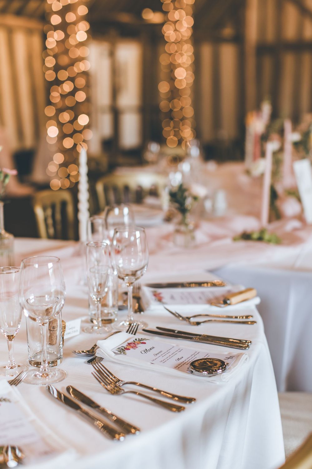 Elegant wedding reception table setting with gold cutlery, crystal glassware, white linens, and warm fairy light bokeh backdrop.