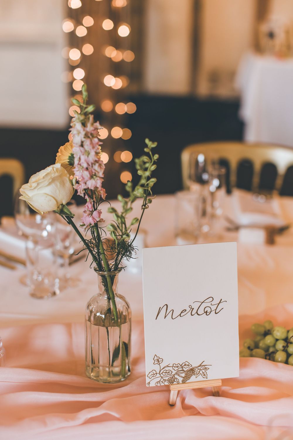 Elegant wedding table setting with a Merlot name card, floral arrangement in glass vase, and bokeh lights in background.