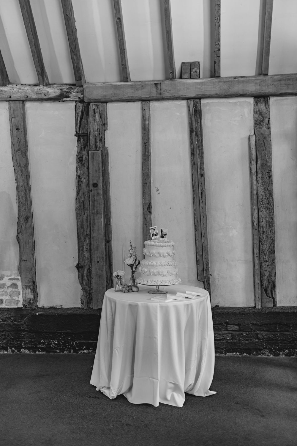 Black and white photo of a tiered wedding cake on a draped table against rustic timber-framed barn walls.