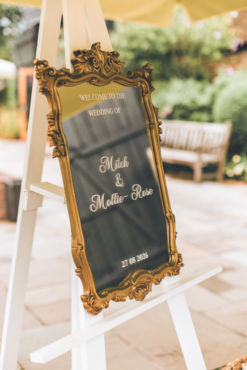 Elegant gold ornate framed wedding welcome sign for Mitch & Mollie-Rose on a white easel outdoors.