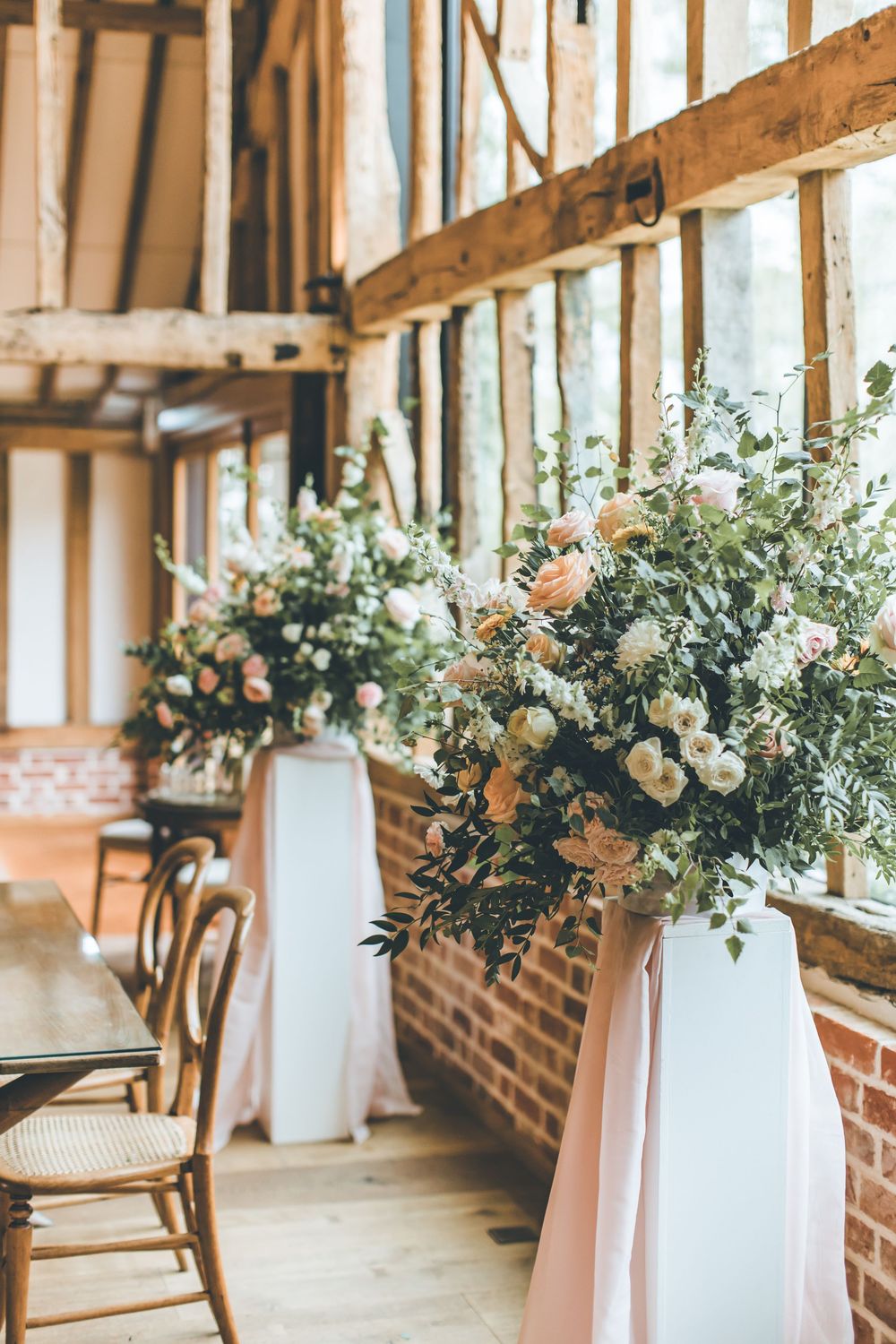 Elegant wedding barn venue with tall floral arrangements of blush roses and greenery on white pedestals with pink draping.