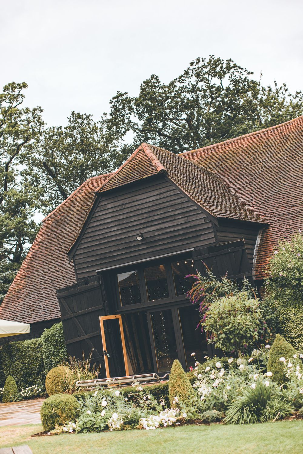 A rustic dark wood barn wedding venue surrounded by lush gardens and mature trees.