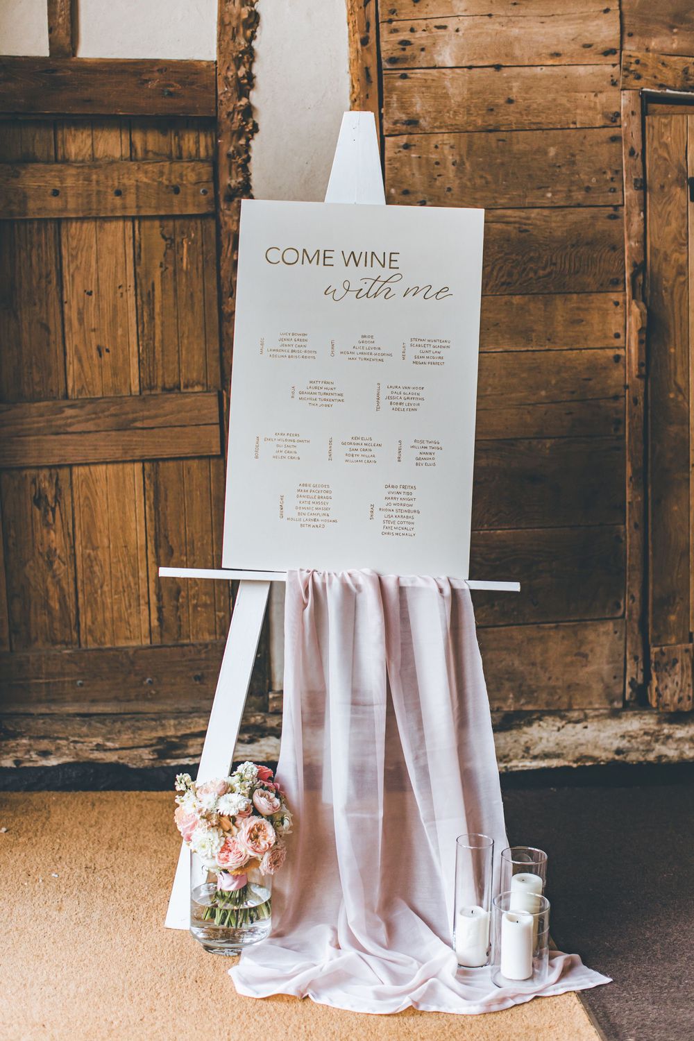 White easel displaying a wedding seating chart with blush fabric draping and floral arrangement in a rustic barn venue.