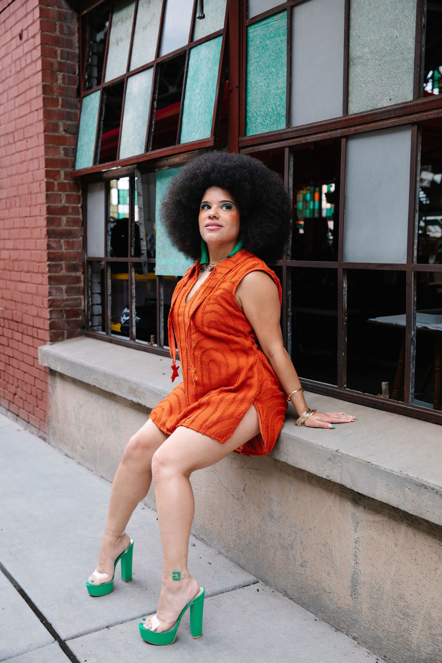 Woman in orange mini dress and green heels with afro hairstyle posing outdoors against brick building.