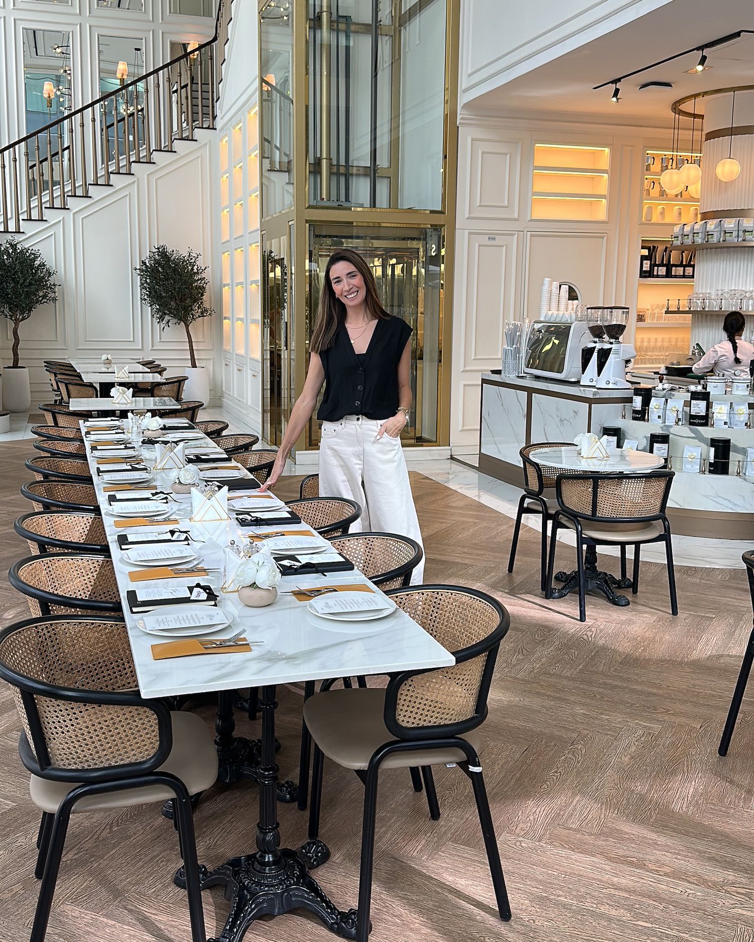 Woman in black top and white pants standing beside a long elegant dining table in a bright upscale restaurant.