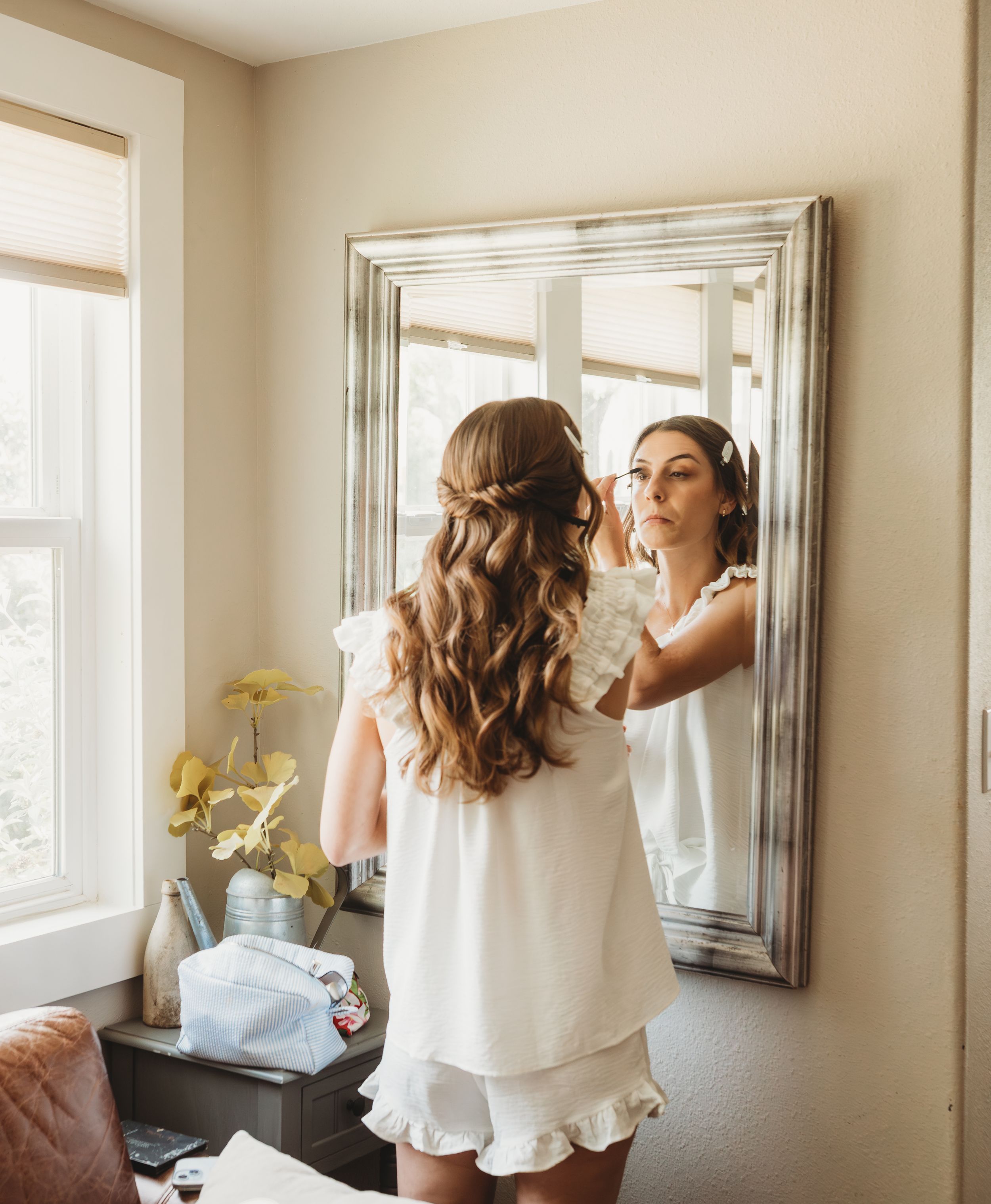 Young girl in white dress with long wavy hair admires herself in a silver-framed mirror while getting ready.
