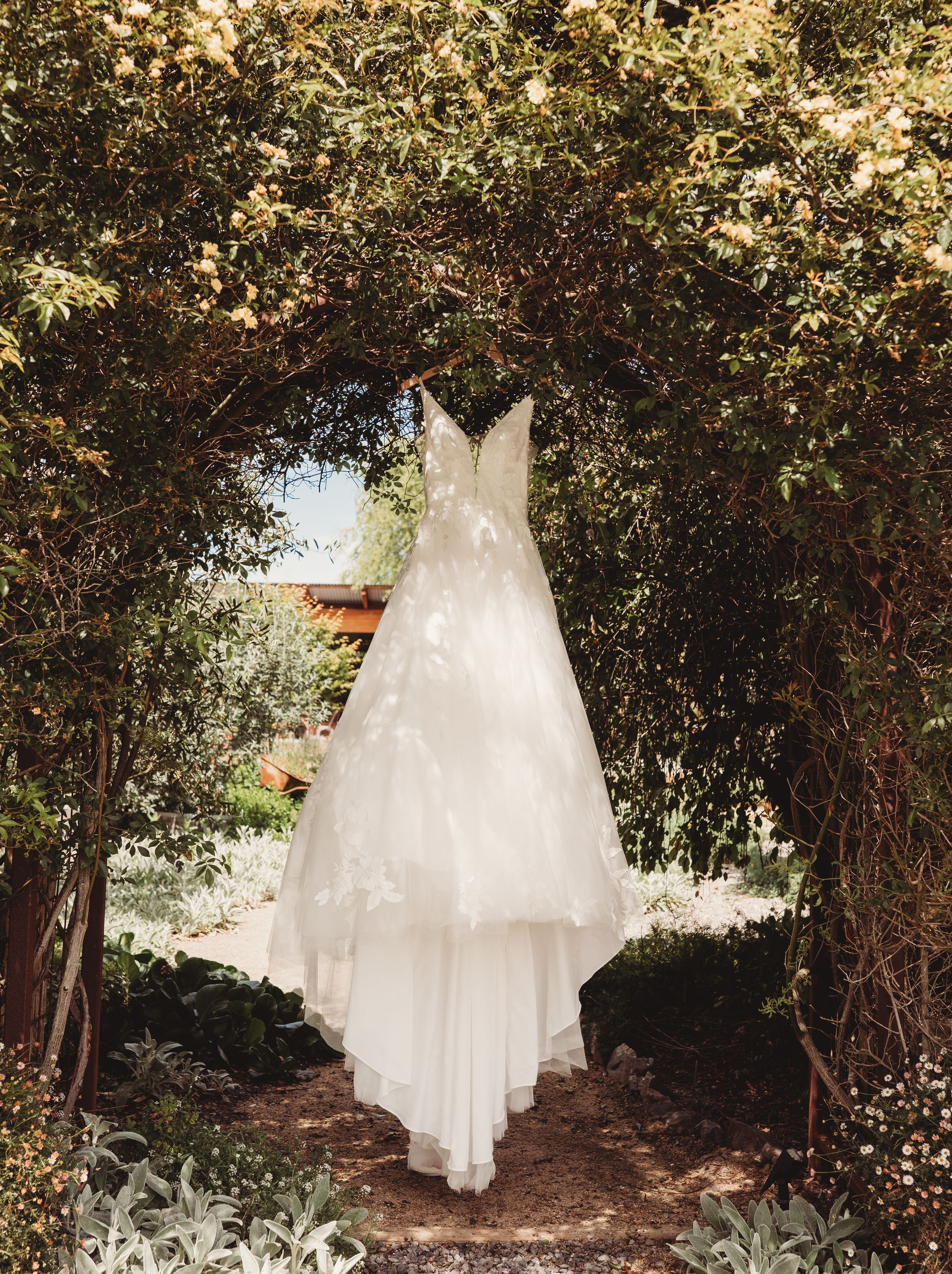 Elegant white wedding gown with floral appliques hanging under a lush green garden archway.