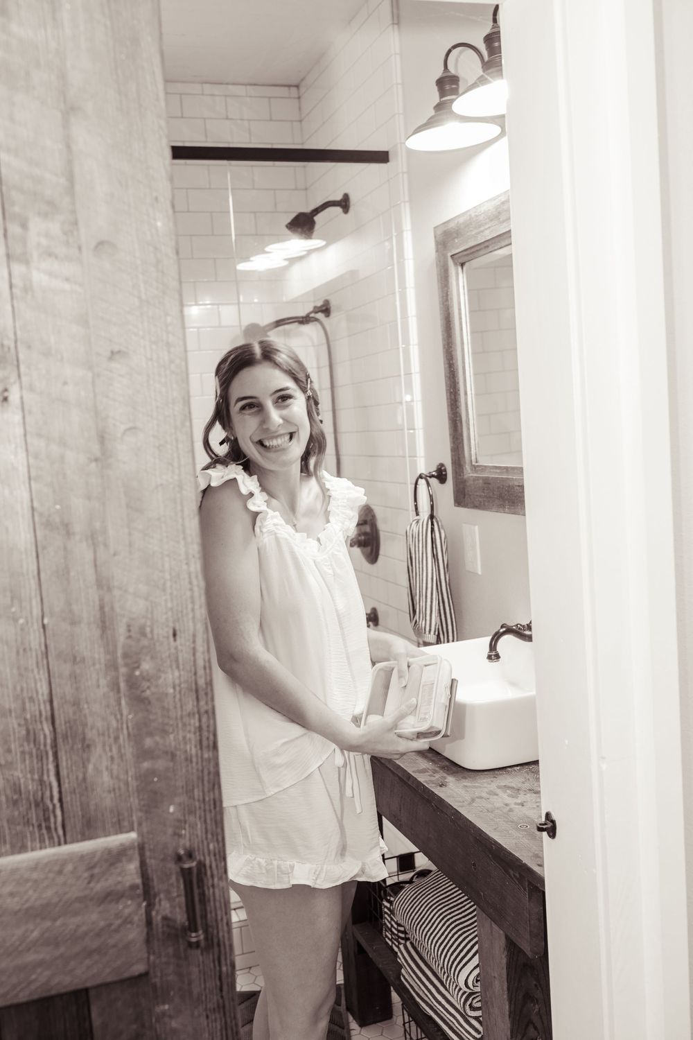 Smiling young woman in white dress holding a book in a rustic farmhouse bathroom with subway tile and wood accents.