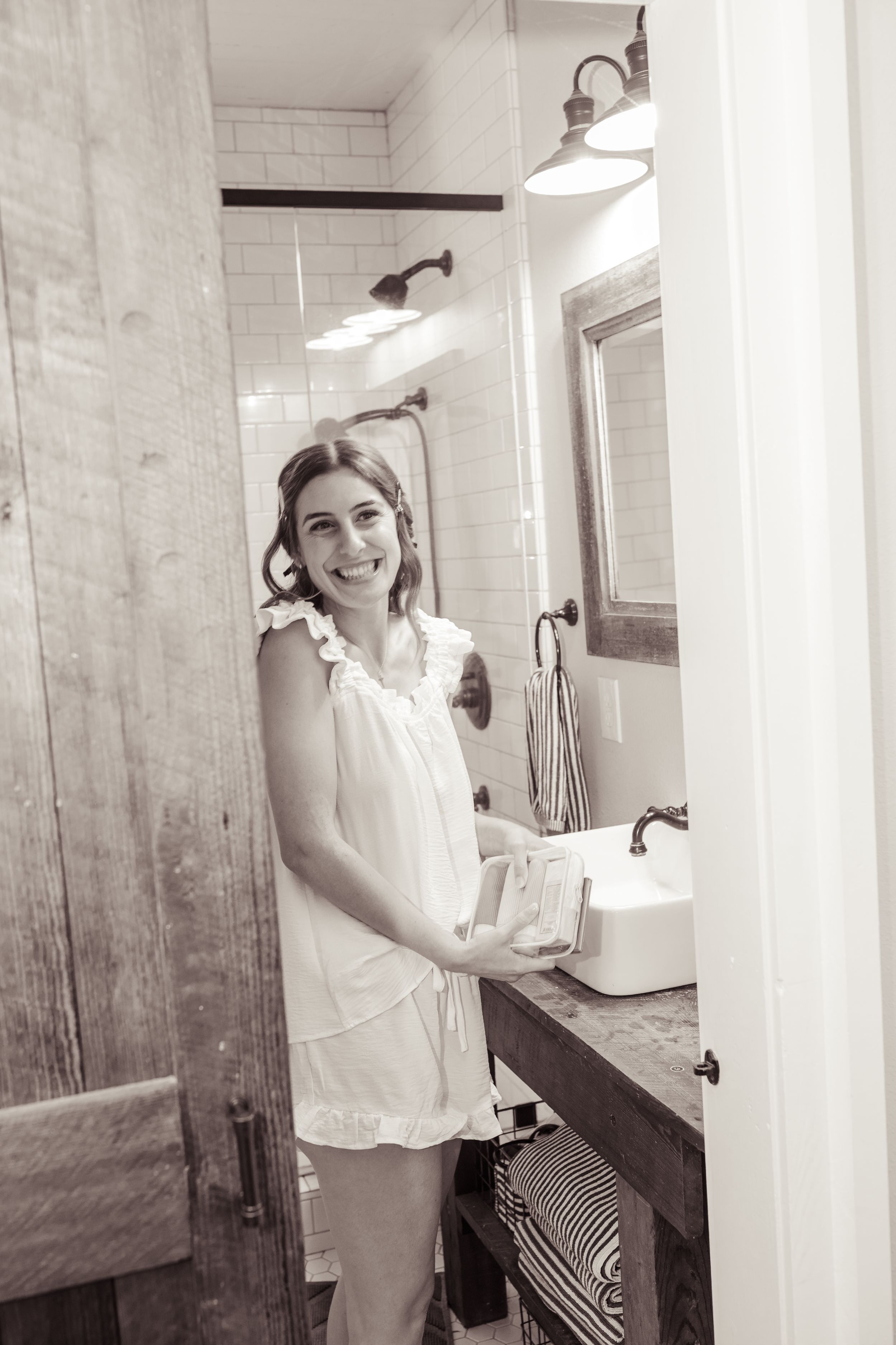 Smiling young woman in white dress holding a book in a rustic farmhouse bathroom with subway tile and wood accents.