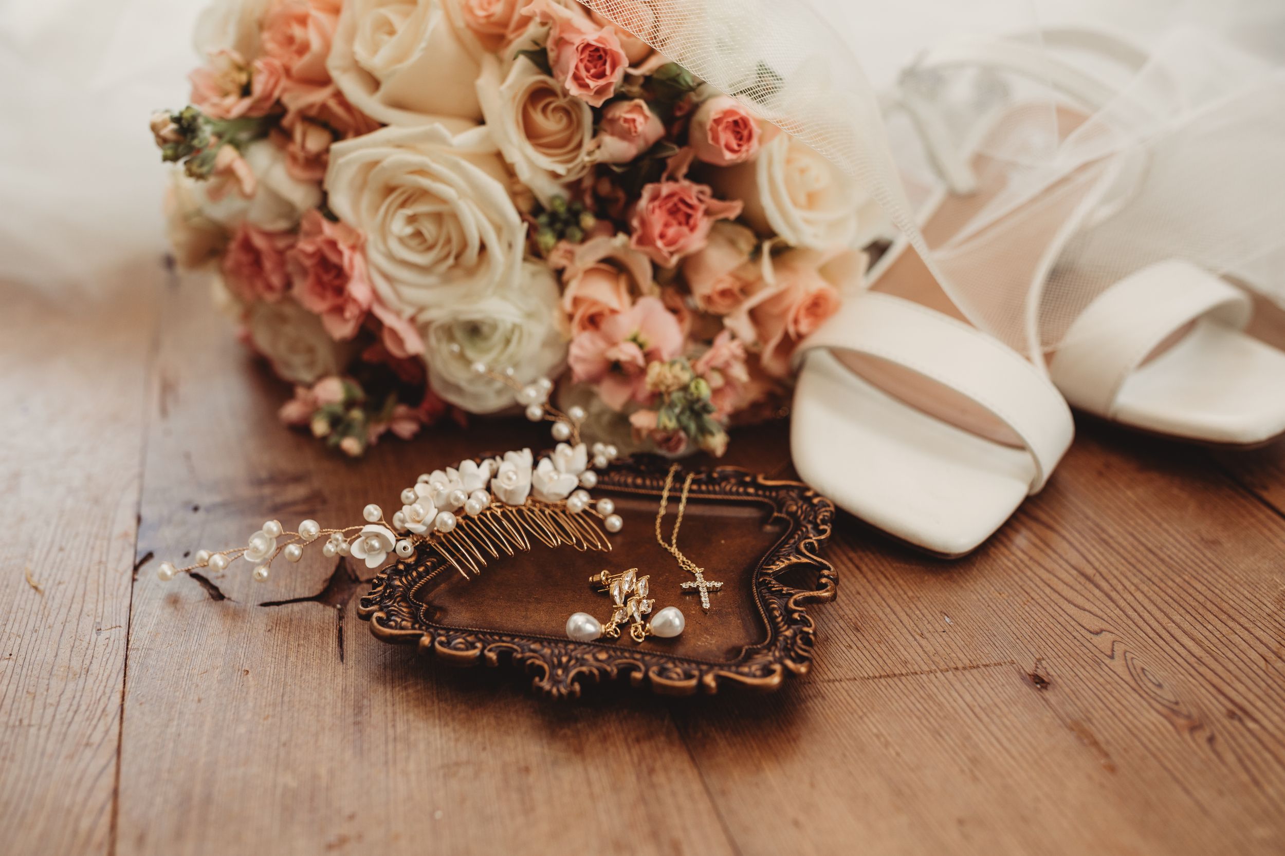 Elegant bridal flat lay with peach roses bouquet, white heels, pearl hair comb, and gold jewelry on wood.