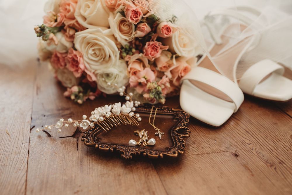 Elegant bridal flat lay with peach roses bouquet, white heels, pearl hair comb, and gold jewelry on wood.