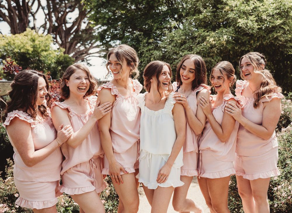Bride and bridesmaids laughing together in matching pink pajama sets in a garden setting.