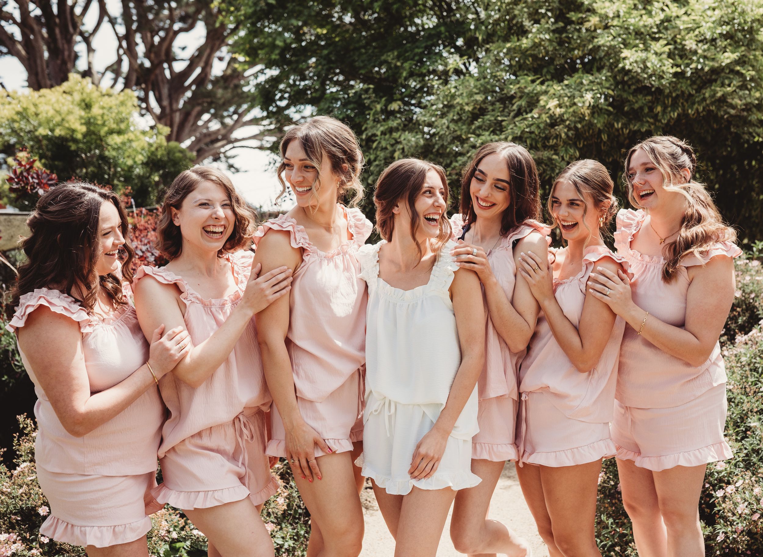 Bride and bridesmaids laughing together in matching pink pajama sets in a garden setting.
