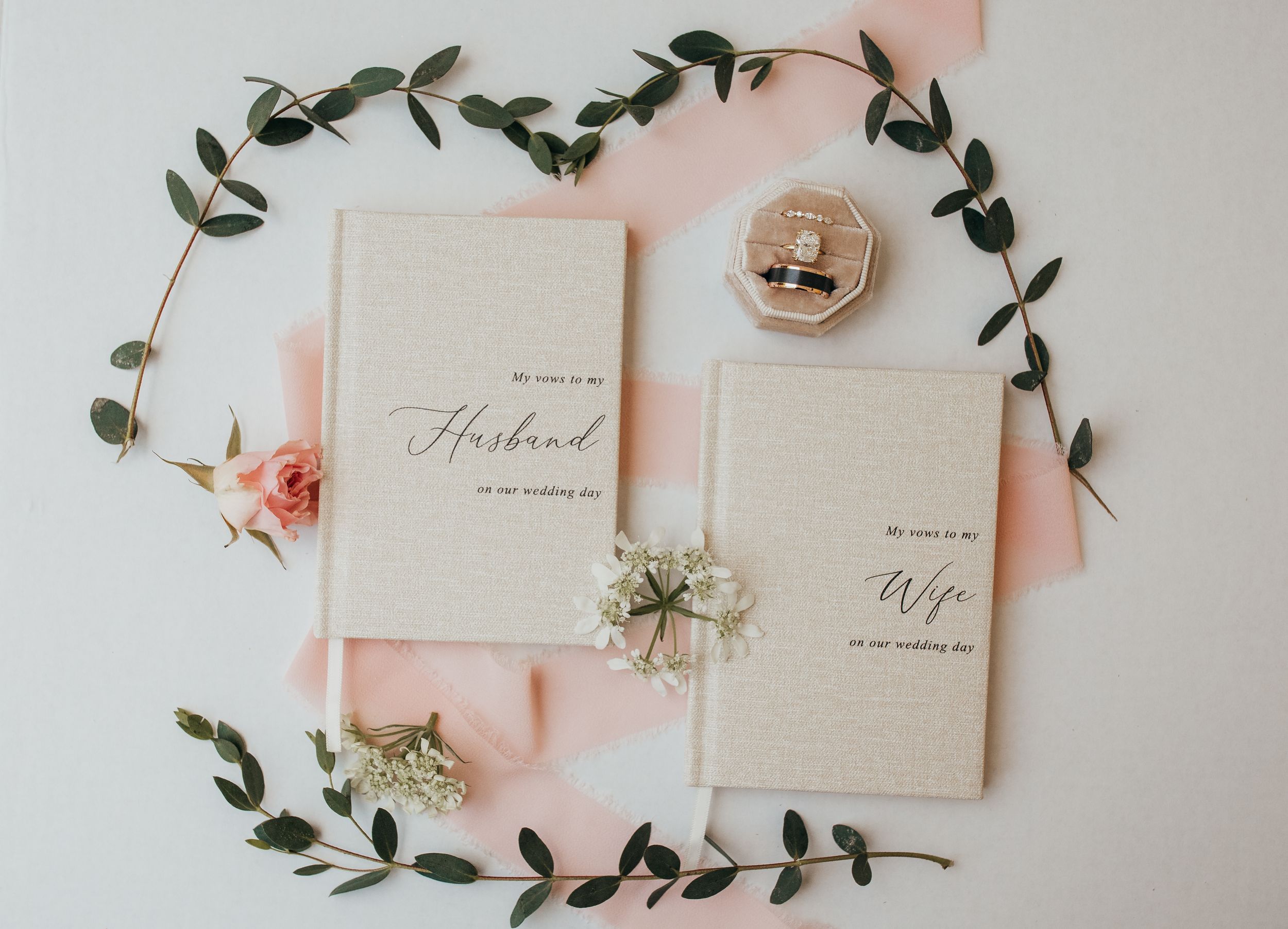 Elegant wedding vow books for husband and wife displayed with floral greenery, pink ribbon, and a velvet ring box.