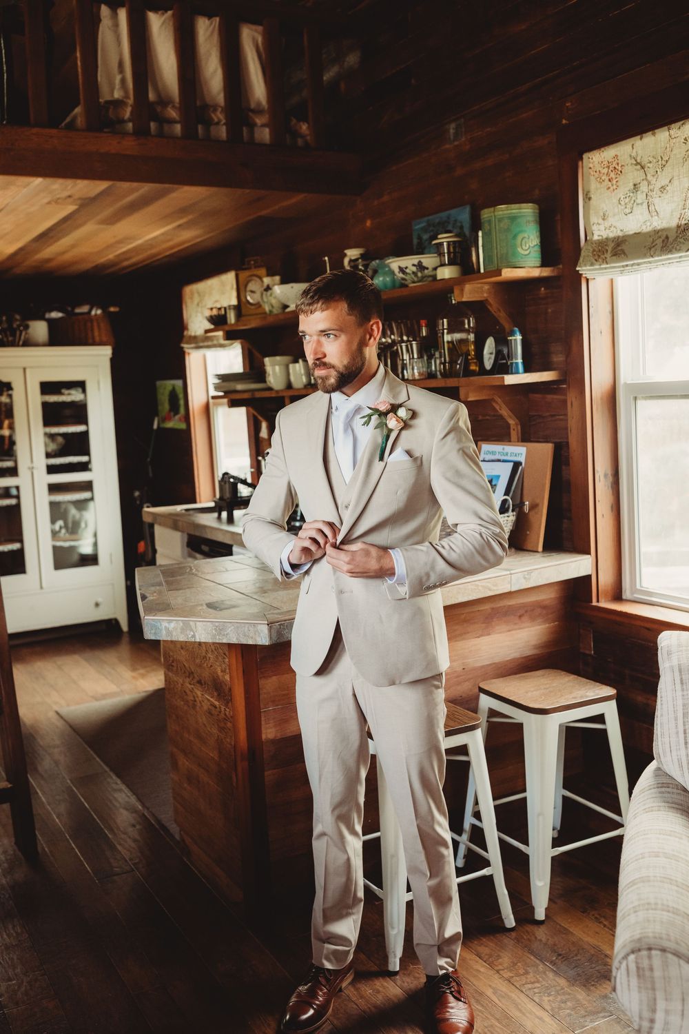 A groom in a beige suit with boutonniere adjusts his jacket in a rustic wood-paneled cabin interior.