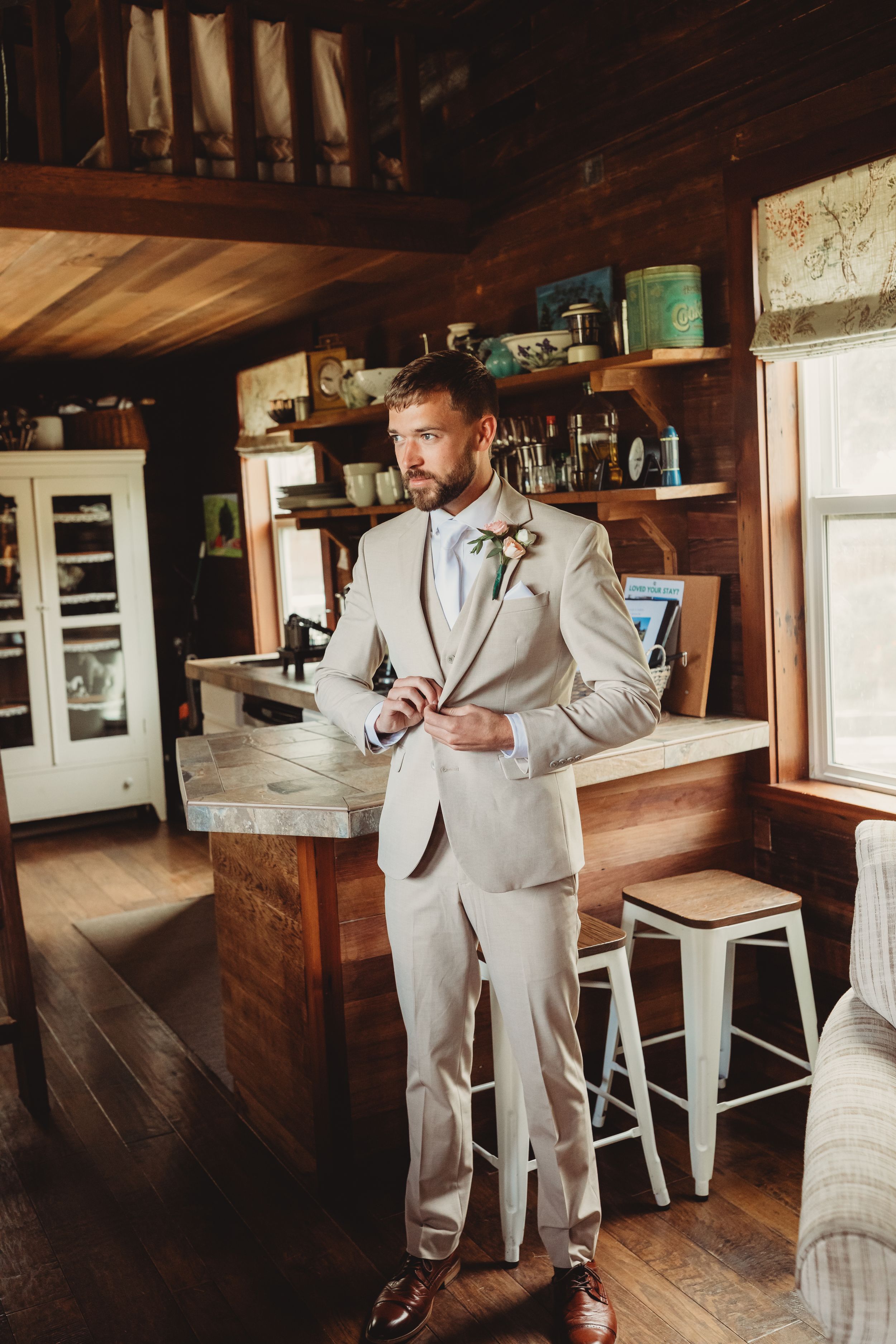 A groom in a beige suit with boutonniere adjusts his jacket in a rustic wood-paneled cabin interior.