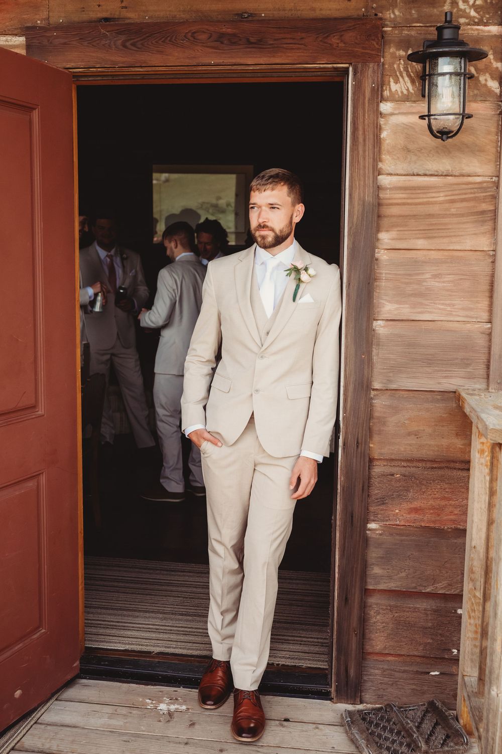 Groom in beige suit with boutonniere stands in rustic barn doorway, groomsmen visible in background.