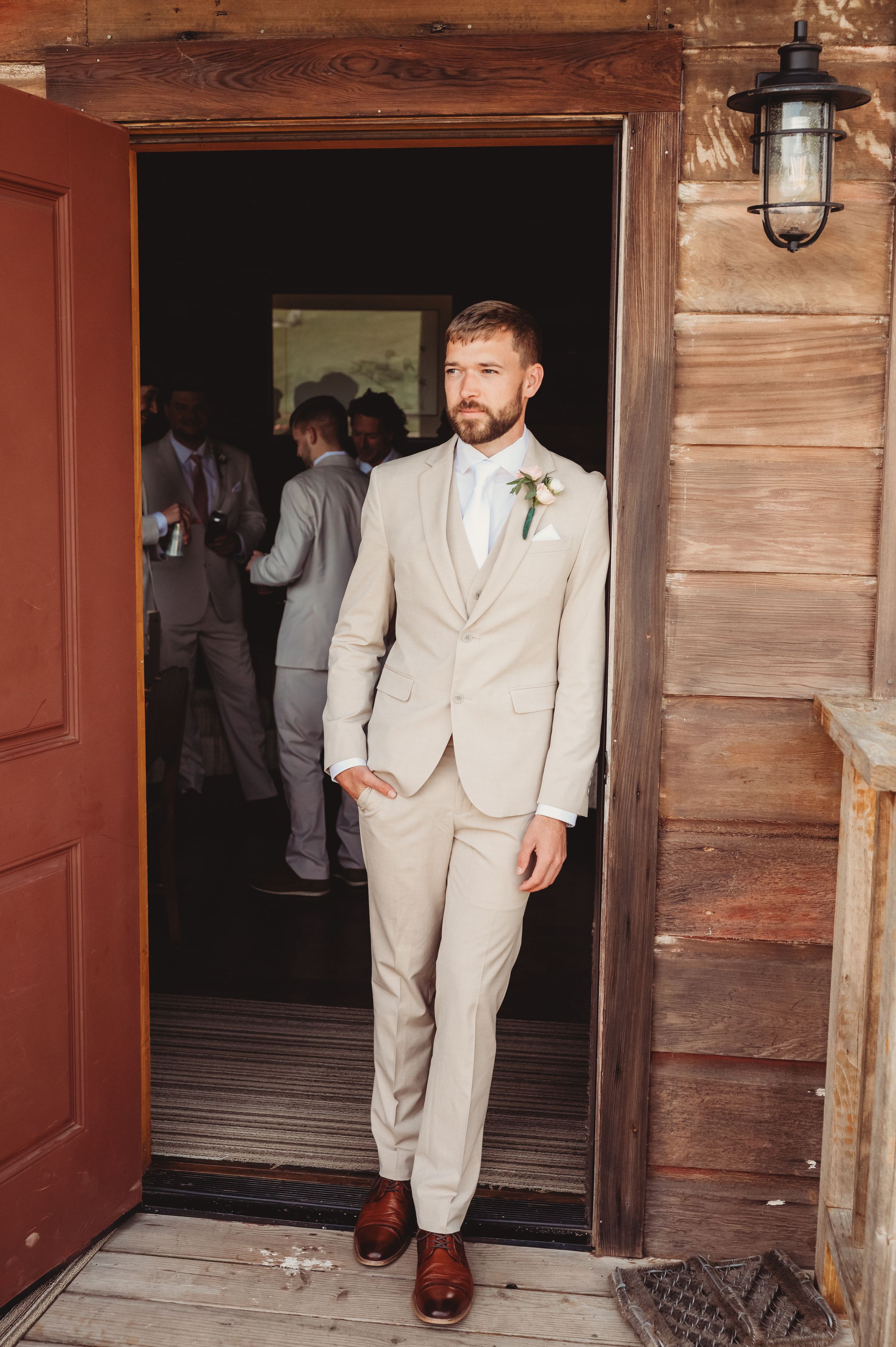 Groom in beige suit with boutonniere stands in rustic barn doorway, groomsmen visible in background.