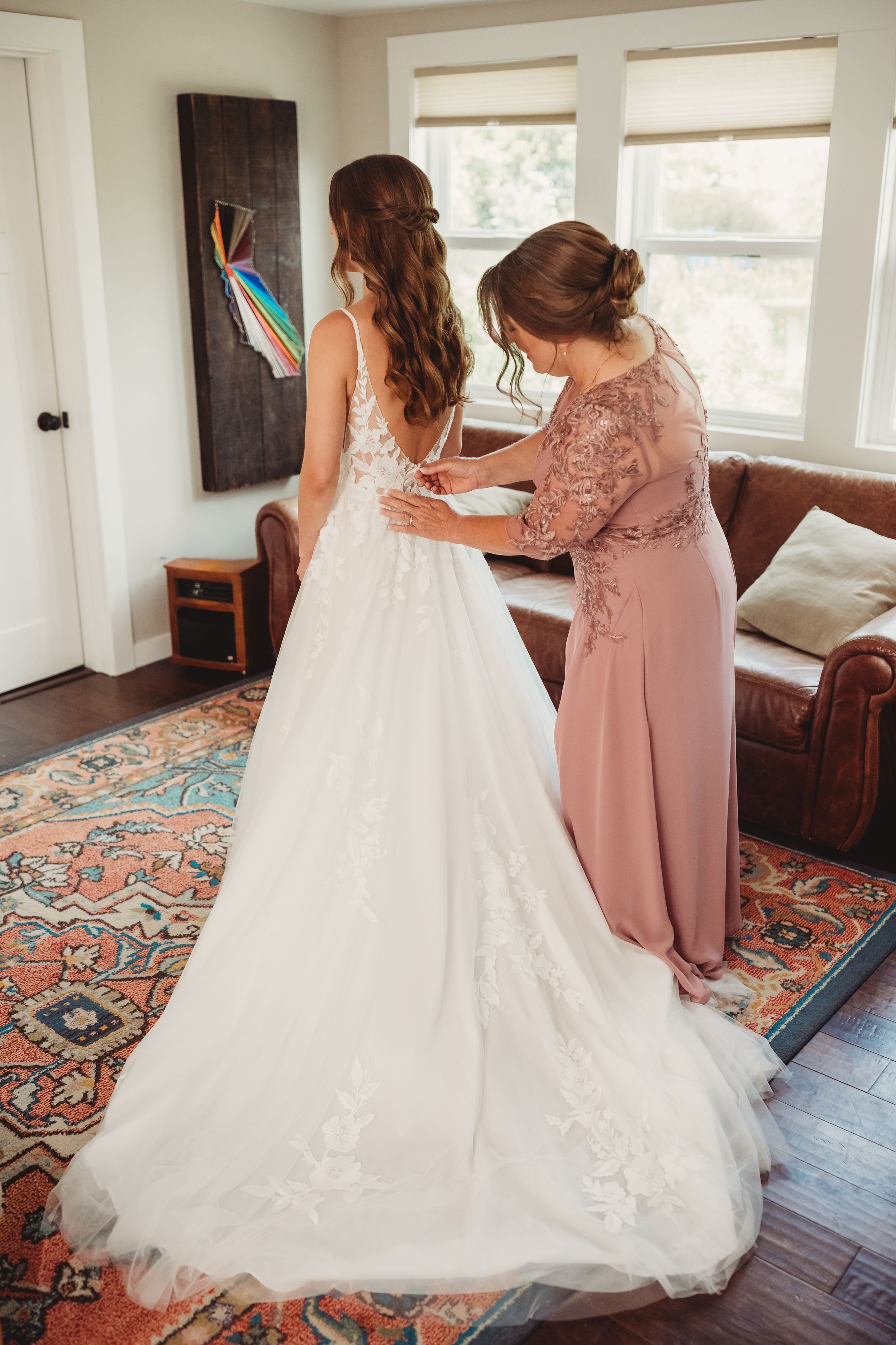 A woman in a blush gown fastens the back of a bride's white lace wedding dress in an elegant living room.