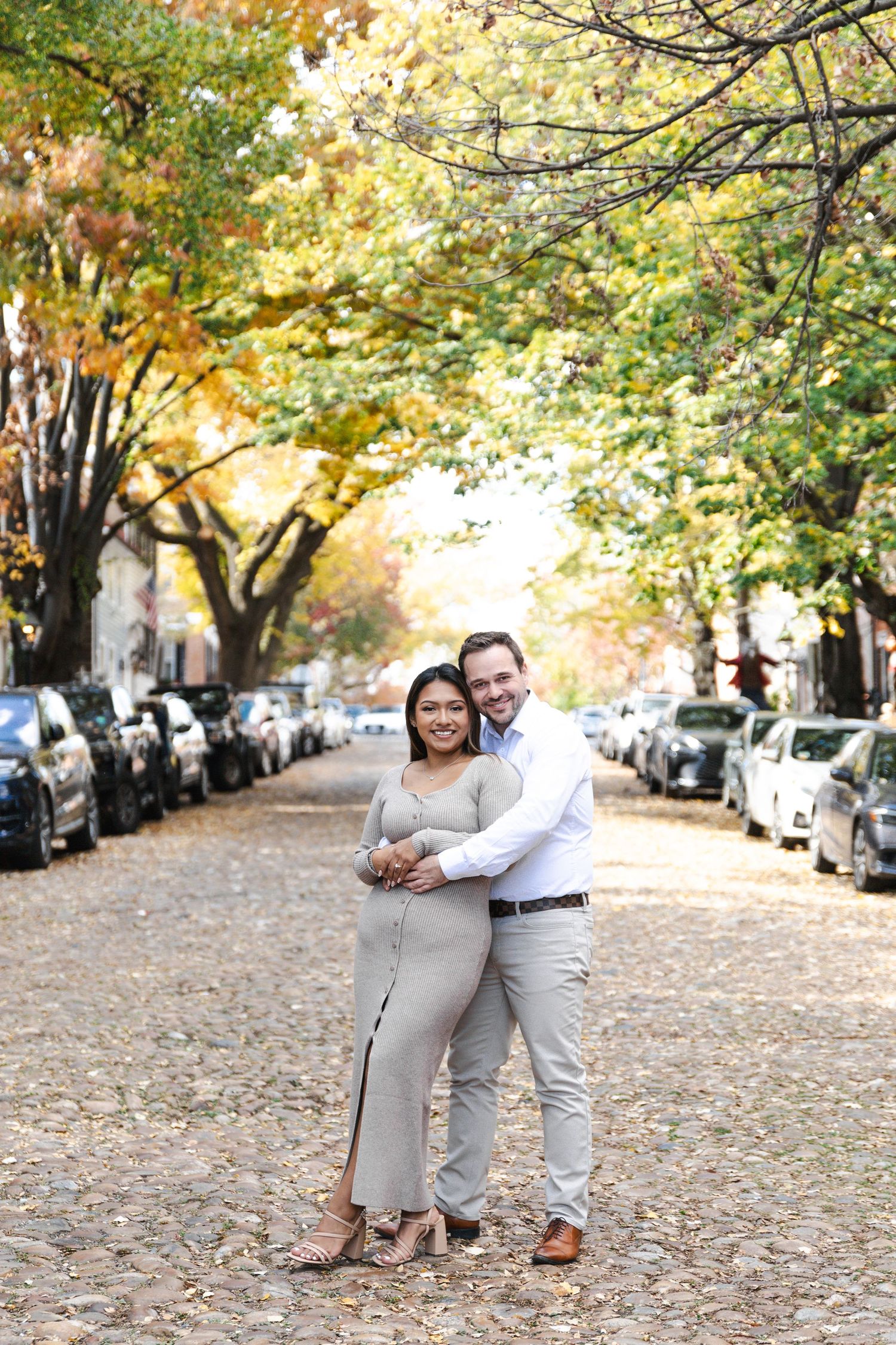 Couple embracing on a cobblestone street lined with autumn trees and parked cars in a charming town.