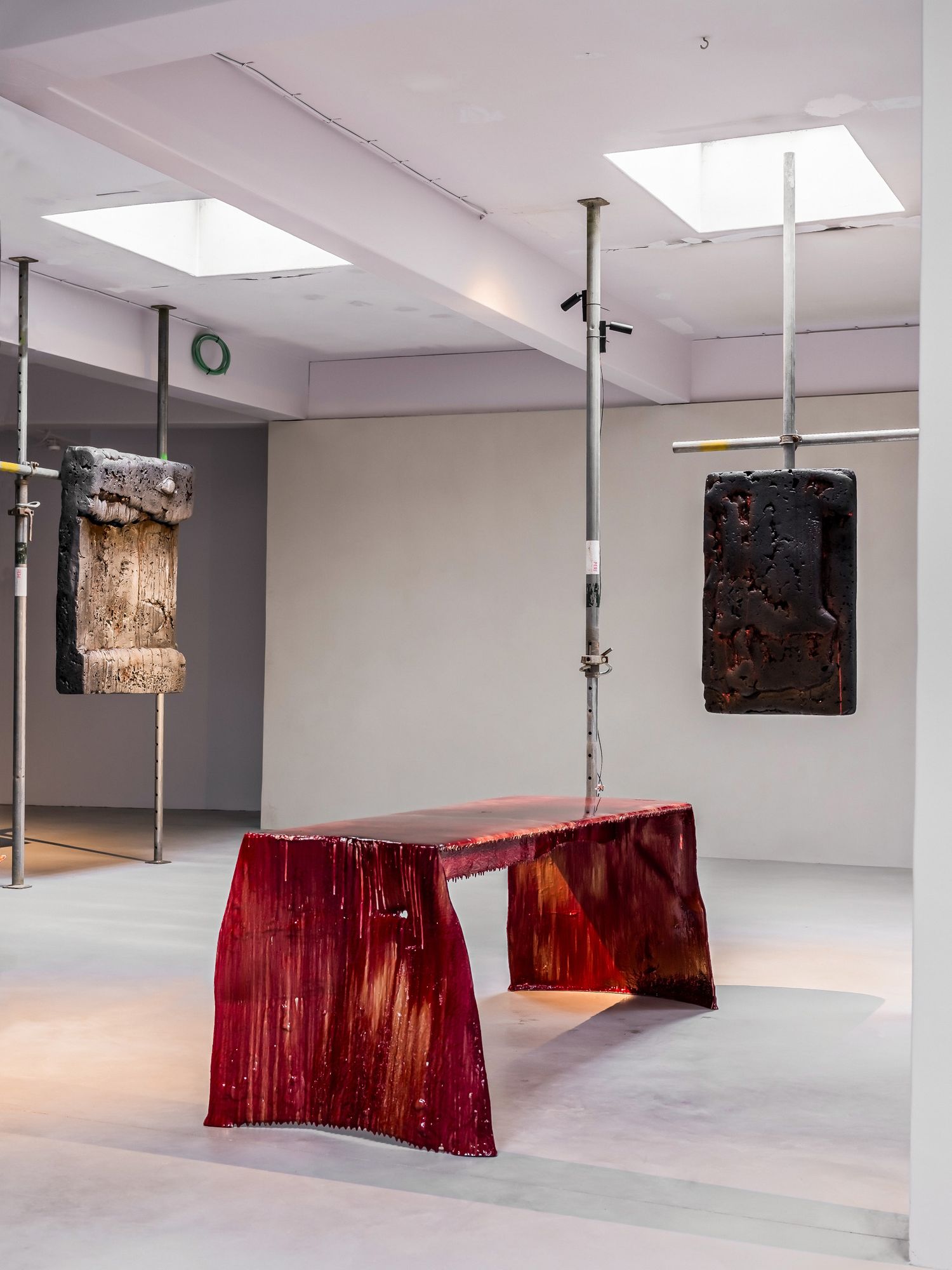 Contemporary art installation featuring a red resin bench, suspended sculptures, and architectural columns in a gallery space.
