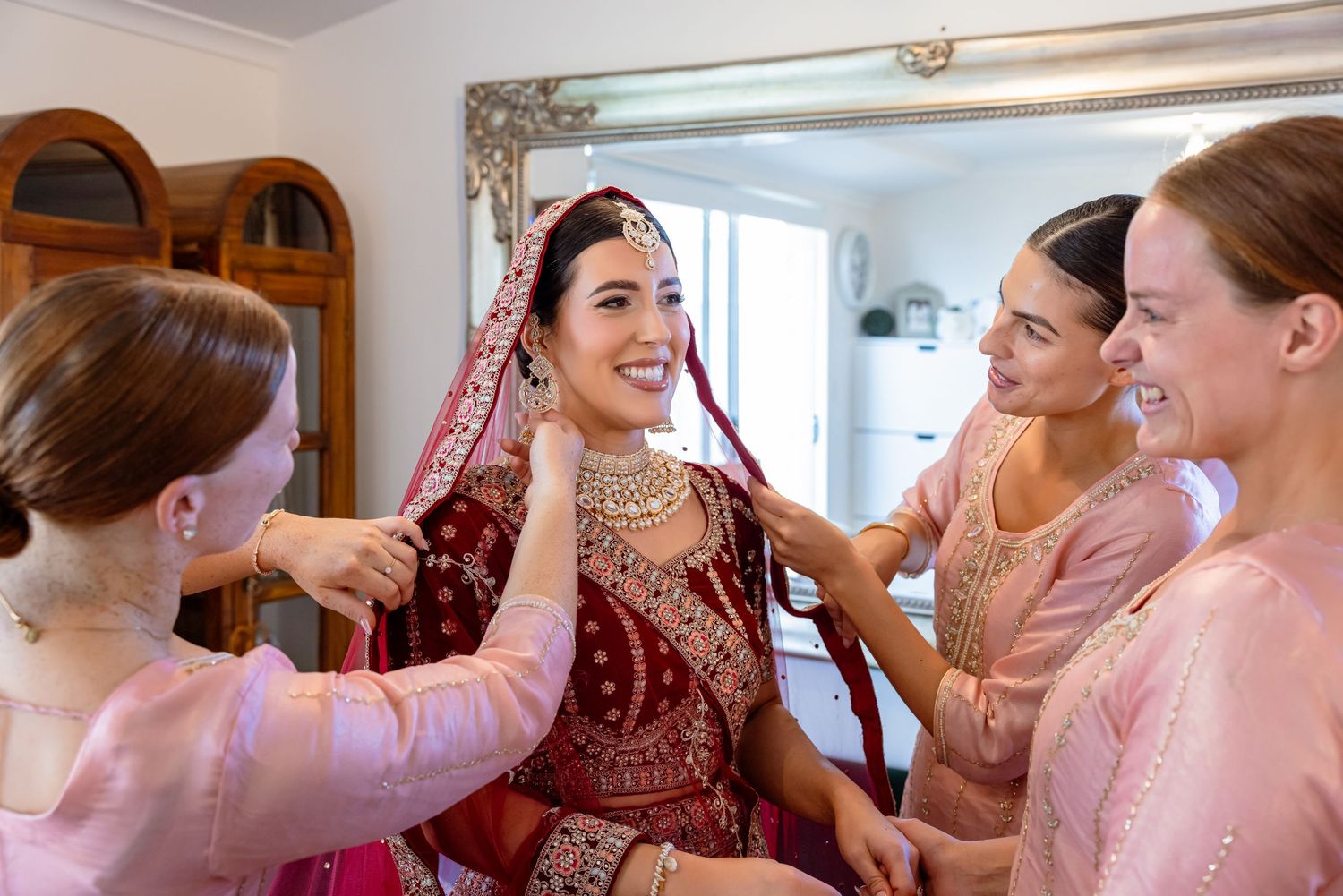 Bride in red bridal lehenga smiling as bridesmaids in pink help her with veil during wedding preparations.