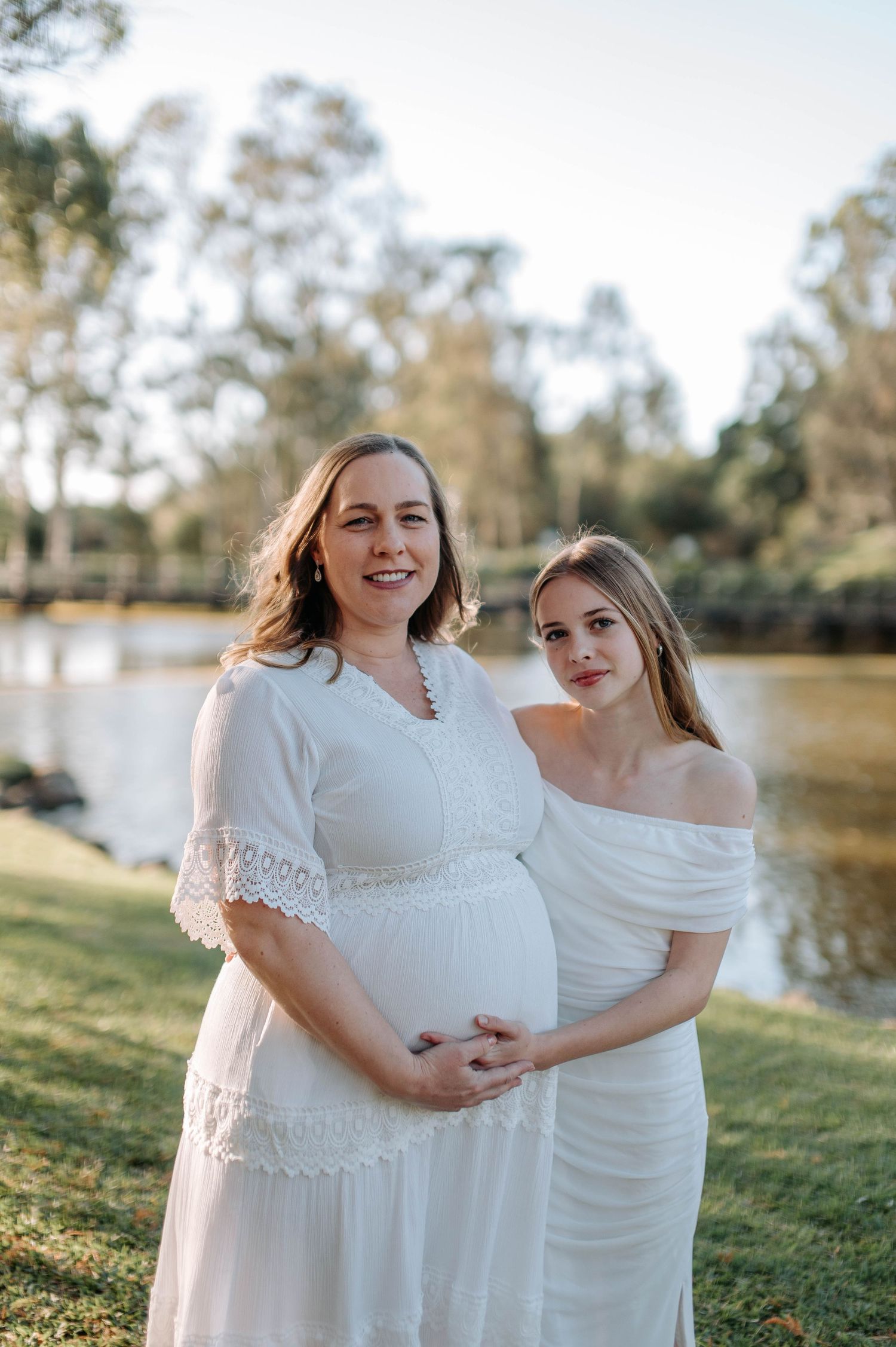 Two pregnant women in white dresses pose together outdoors near a serene river surrounded by lush green trees.