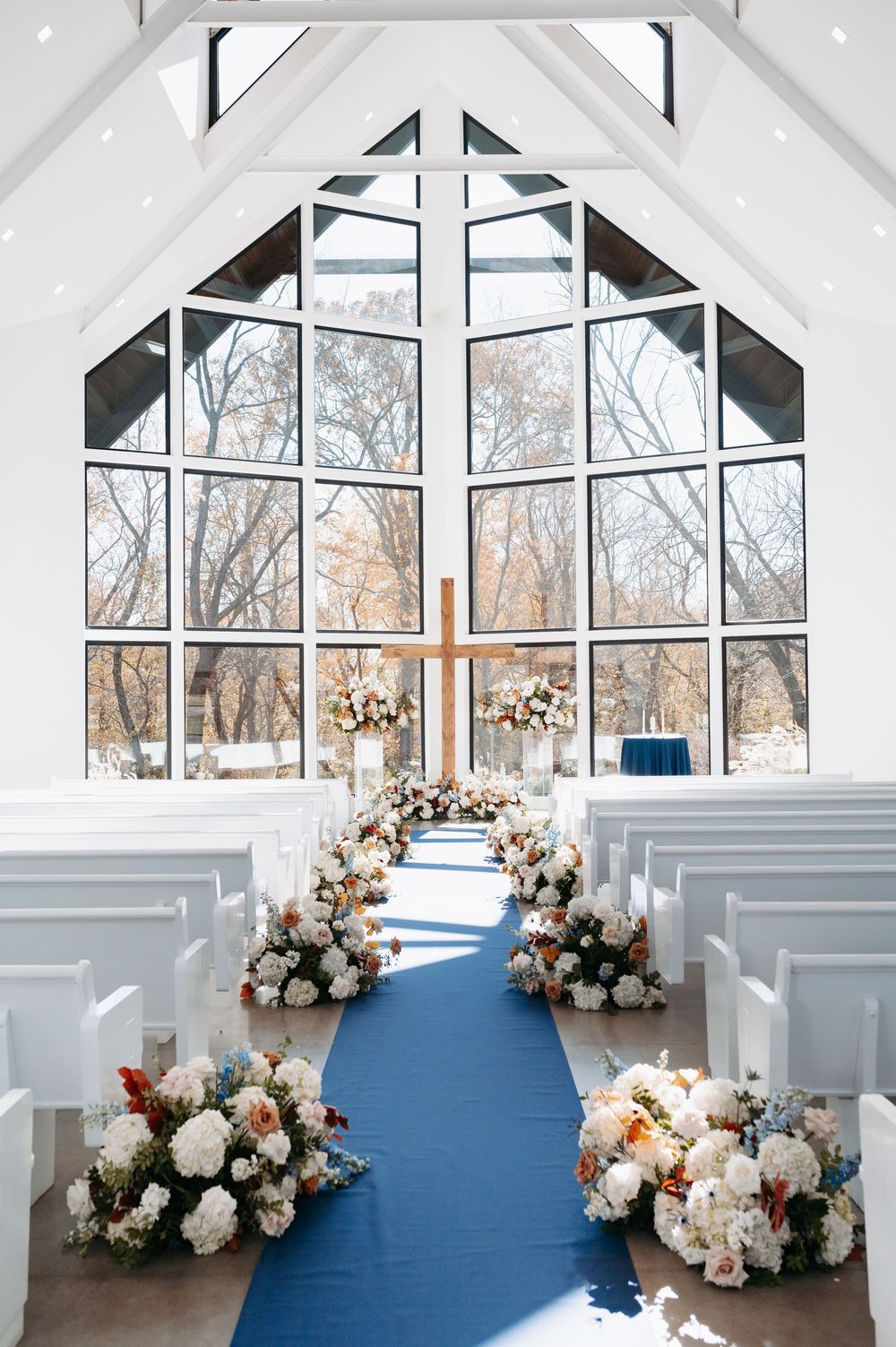A bright white chapel wedding aisle with a blue runner, white pews, and lush floral arrangements leading to a wooden cross.