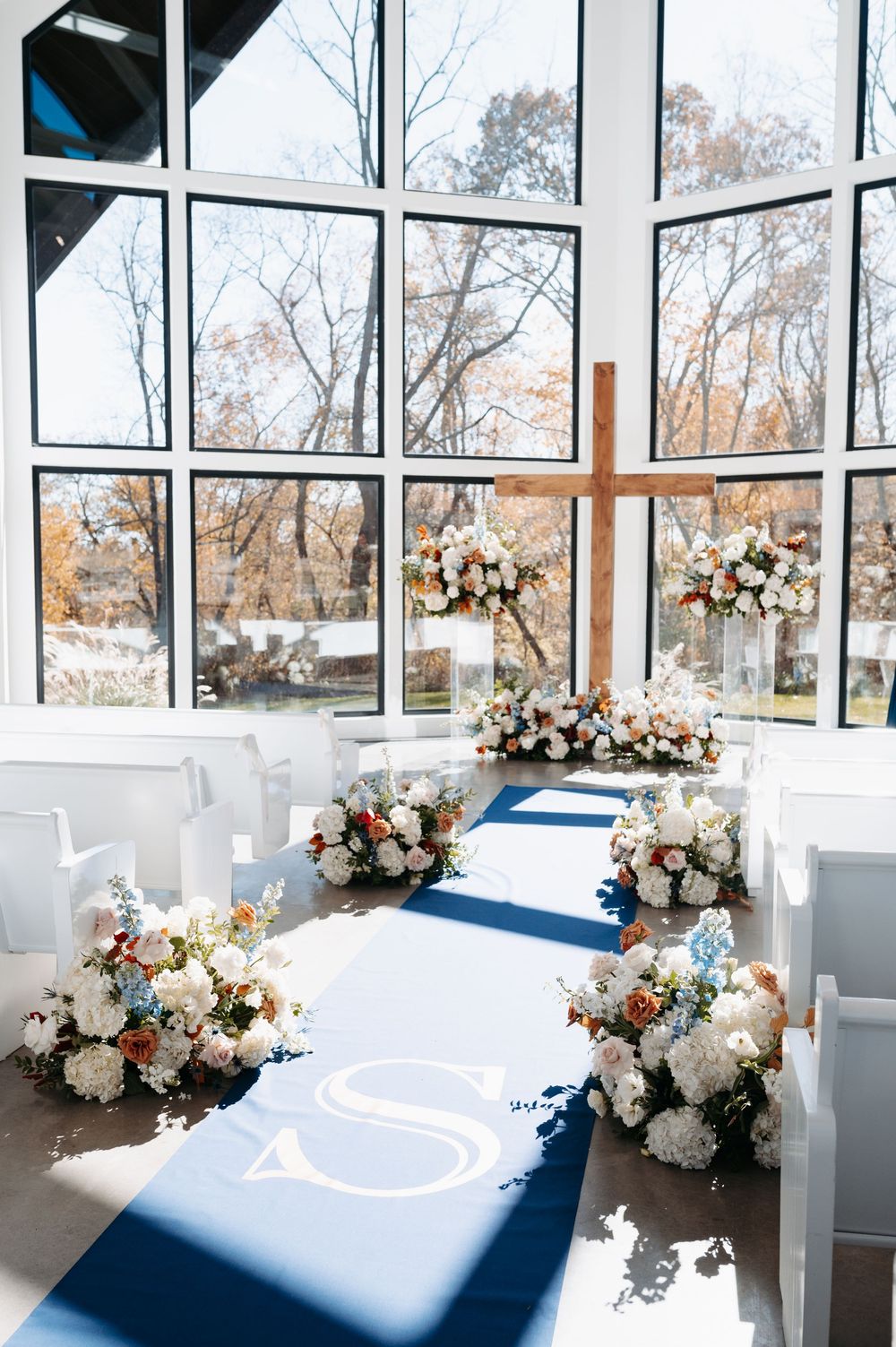 Elegant wedding chapel aisle with navy runner, monogram, white floral arrangements, and wooden cross.