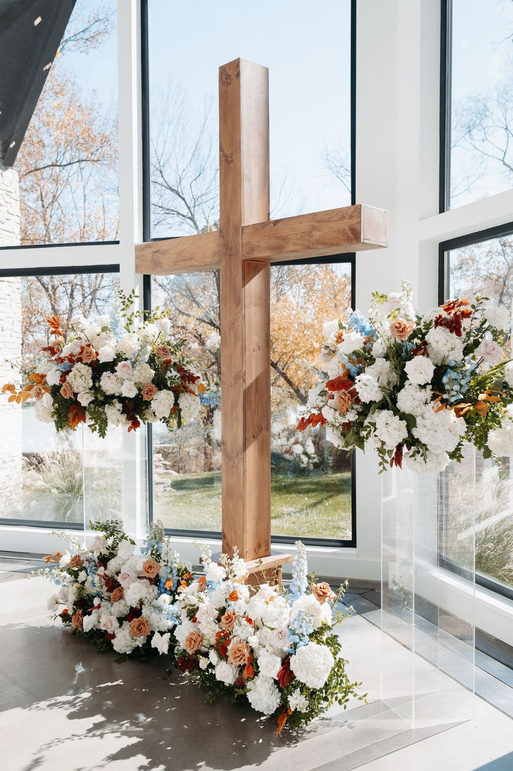 A rustic wooden cross adorned with white, orange, and blue floral arrangements in a sunlit chapel with large windows.
