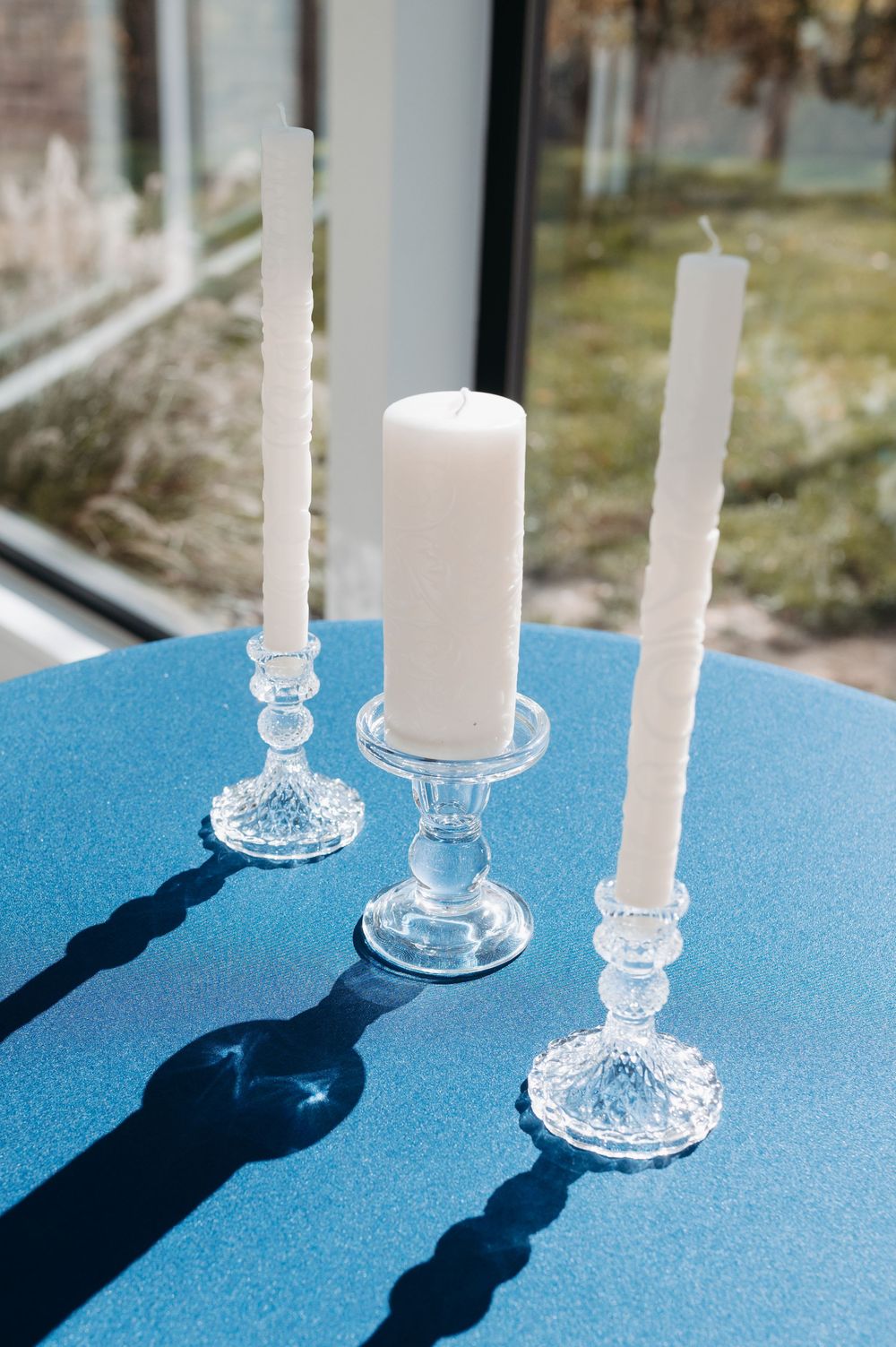 Three white candles in crystal glass holders casting shadows on a blue tablecloth near a sunlit window.