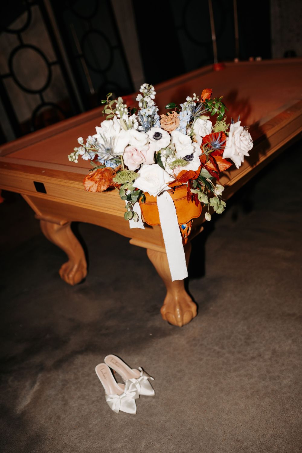 Bridal bouquet with white, blush, and orange blooms resting on a wooden billiard table beside satin heels.