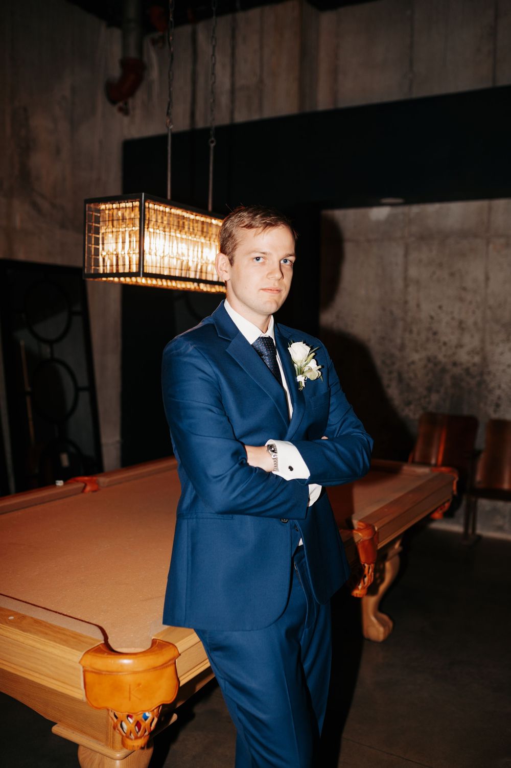 Groom in navy blue suit with boutonniere poses confidently beside a pool table in an elegant venue.