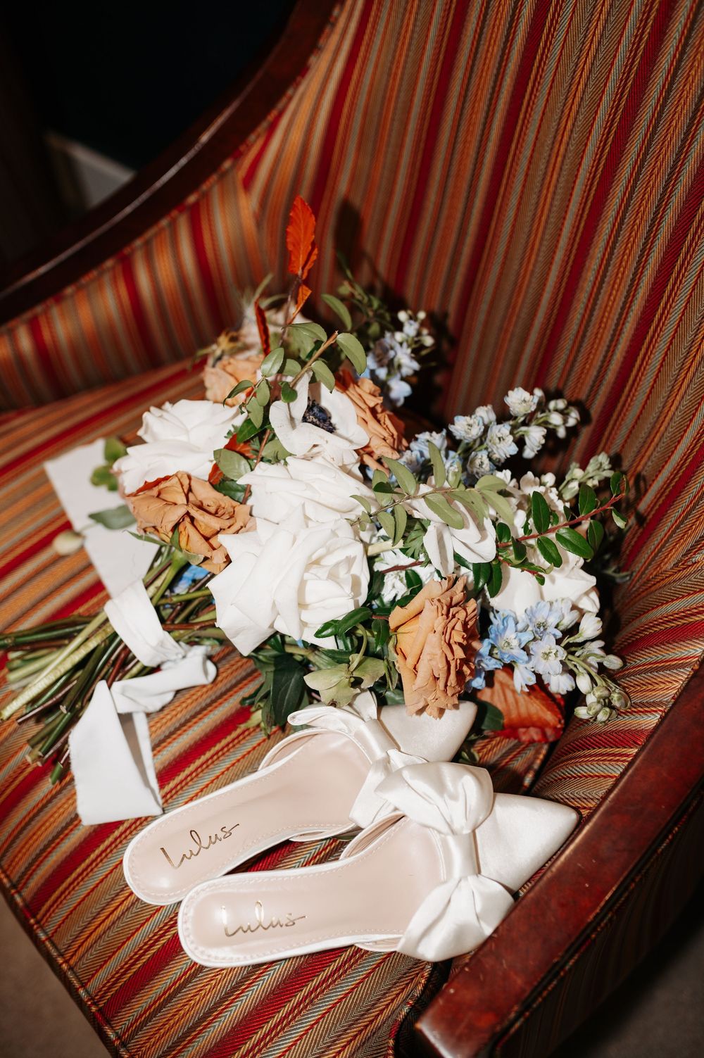 White bow heels by Lulus beside a bridal bouquet of orange, white, and blue flowers on a striped chair.