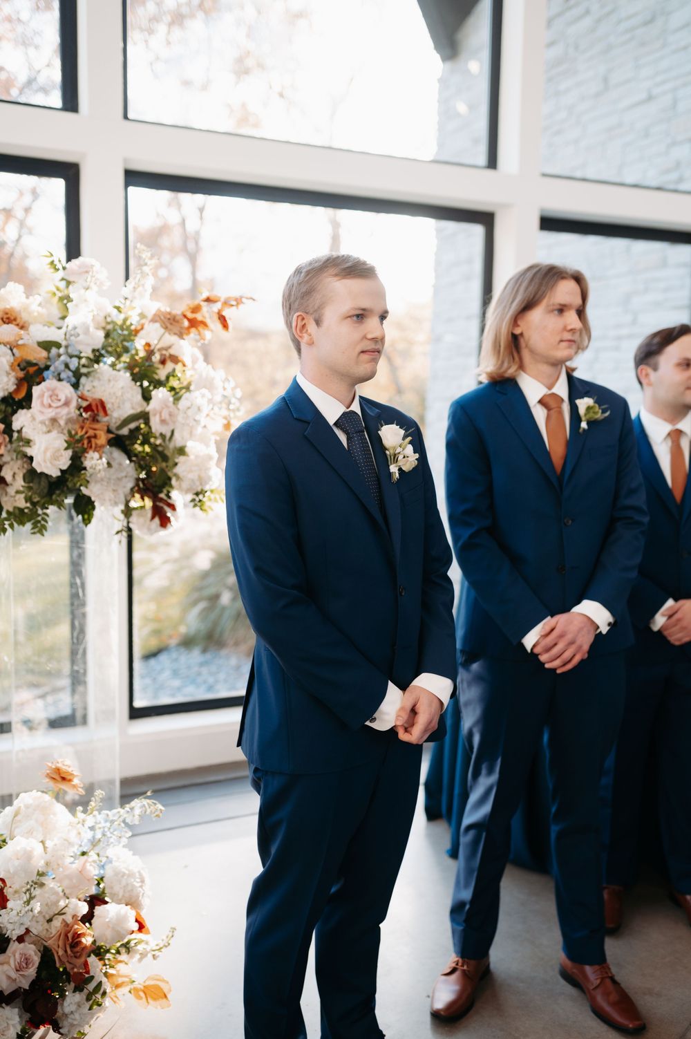 Groom in navy blue suit standing at wedding altar with floral arrangements and groomsmen in background.