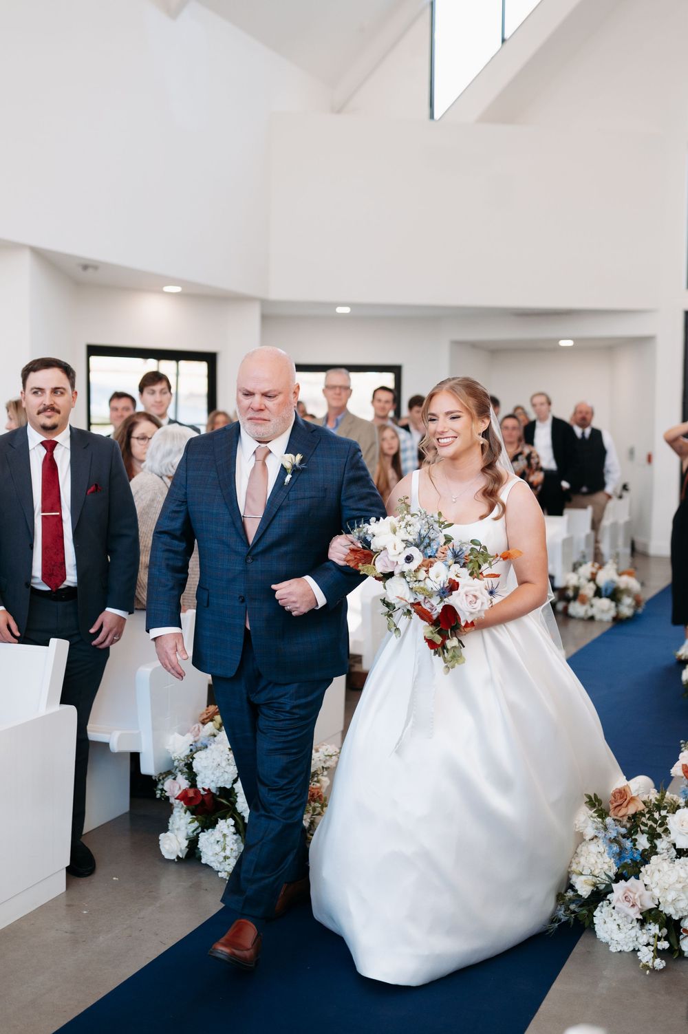Bride in white gown walks down blue aisle with escort holding red and white bouquet at indoor wedding ceremony.