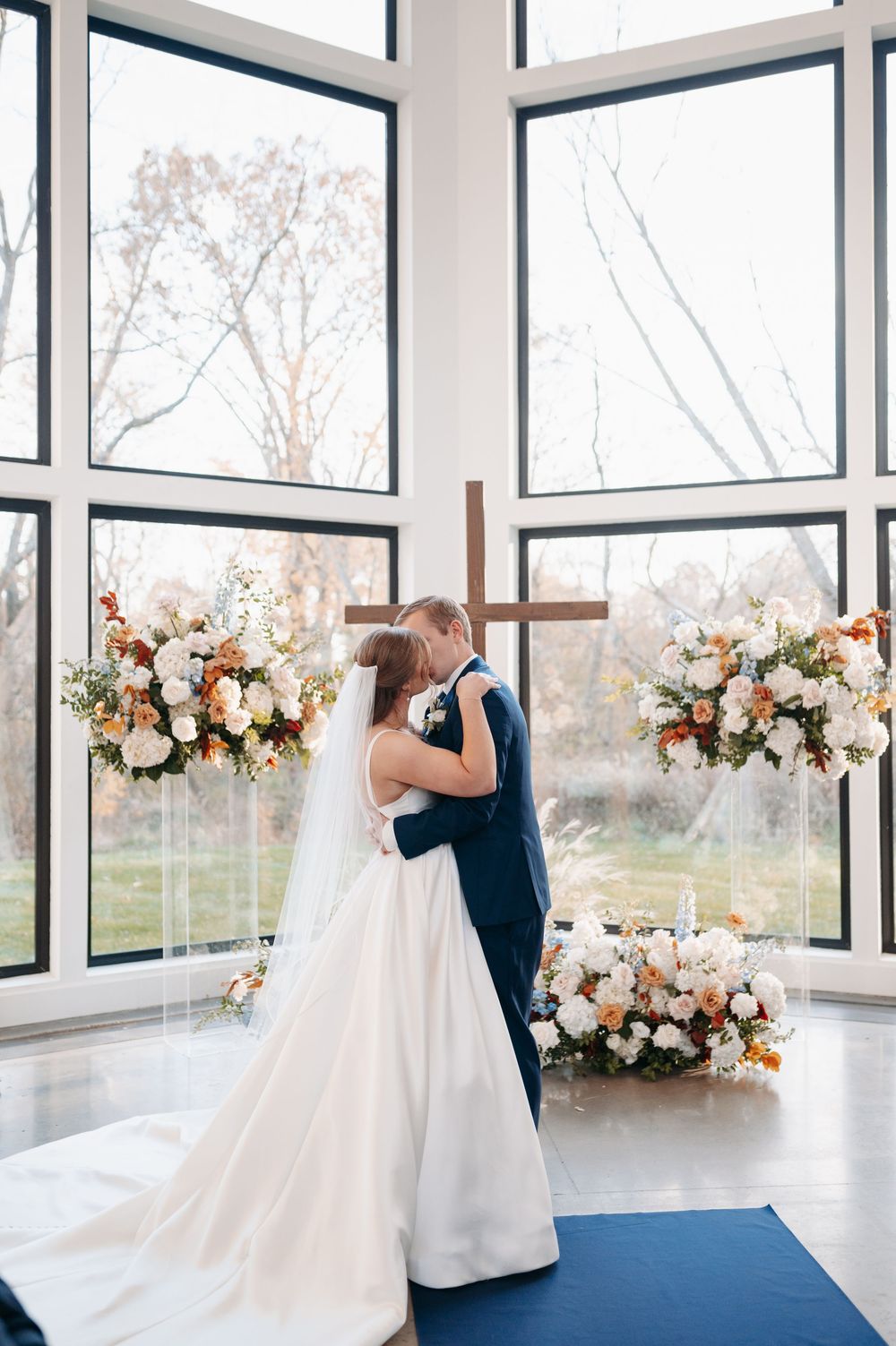 Bride and groom share first kiss at altar with wooden cross and elegant floral arrangements in a sunlit chapel.