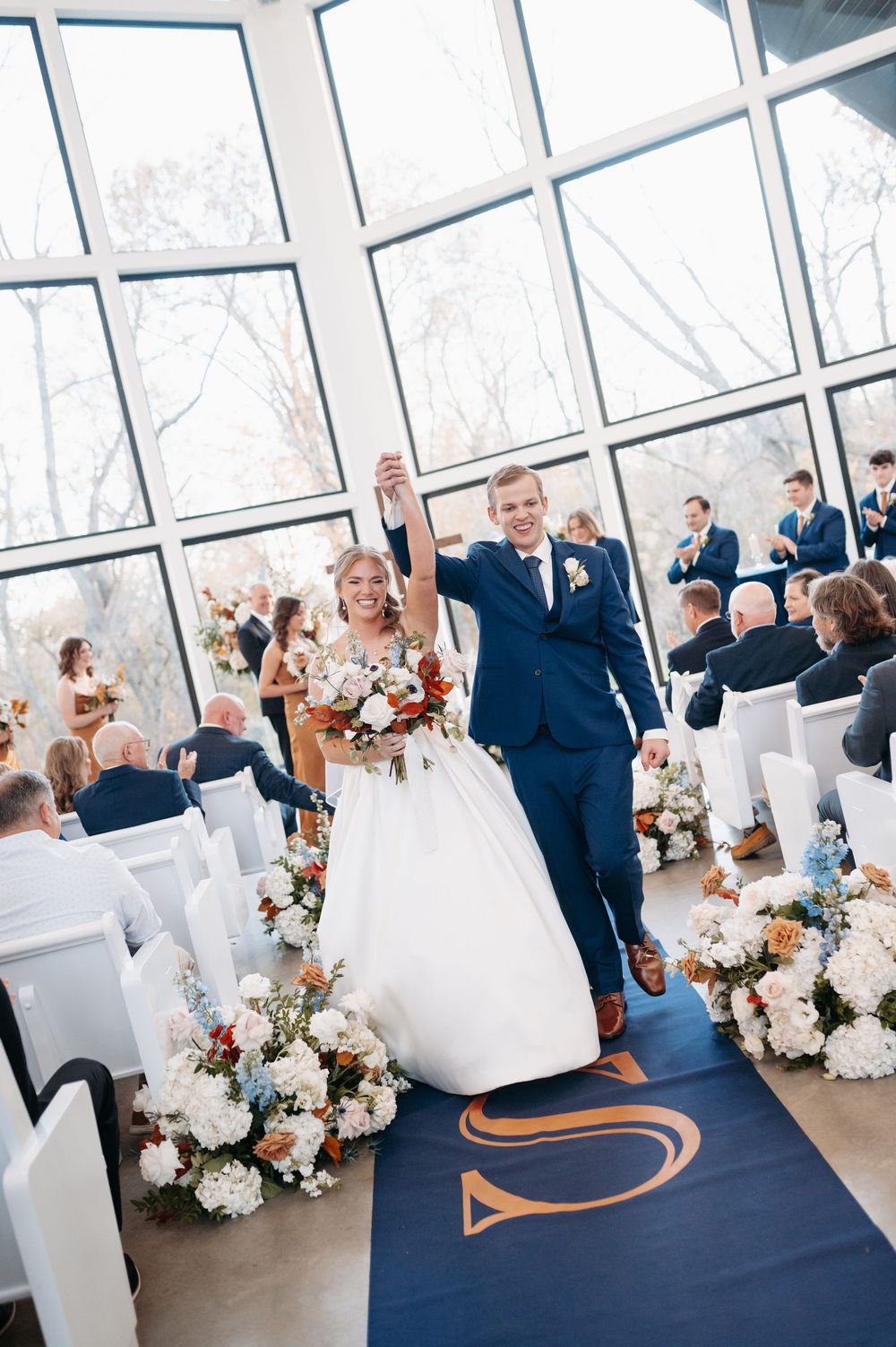 Bride and groom raise hands in joy walking down aisle in sunlit chapel with floral arrangements and guests.