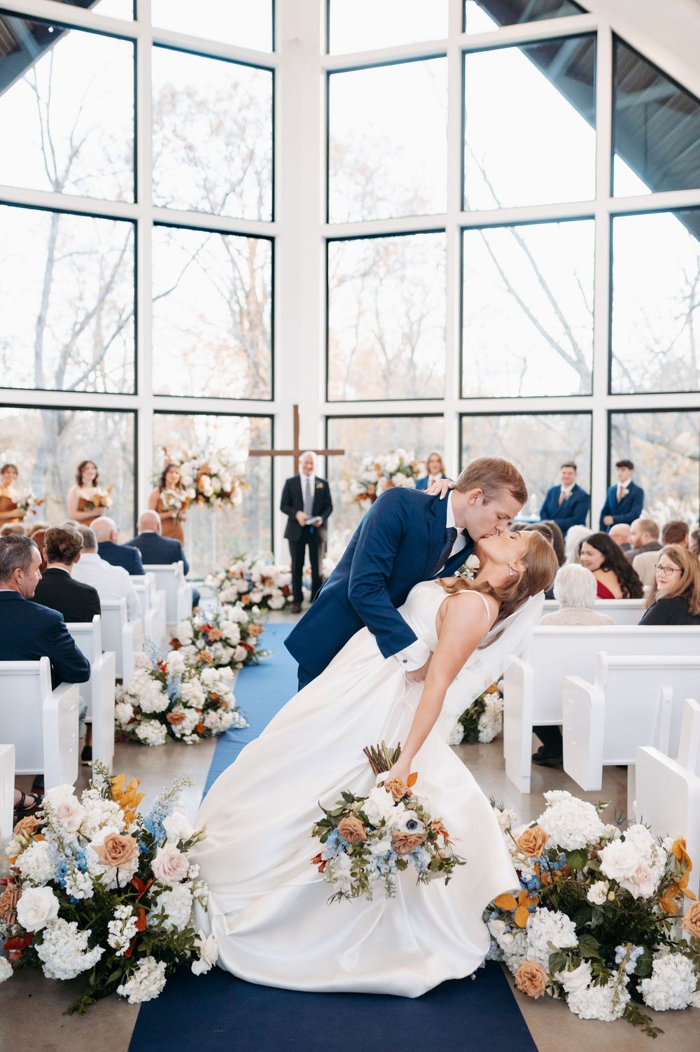 Groom dips bride for a kiss at wedding ceremony altar in a modern chapel with large windows and floral arrangements.