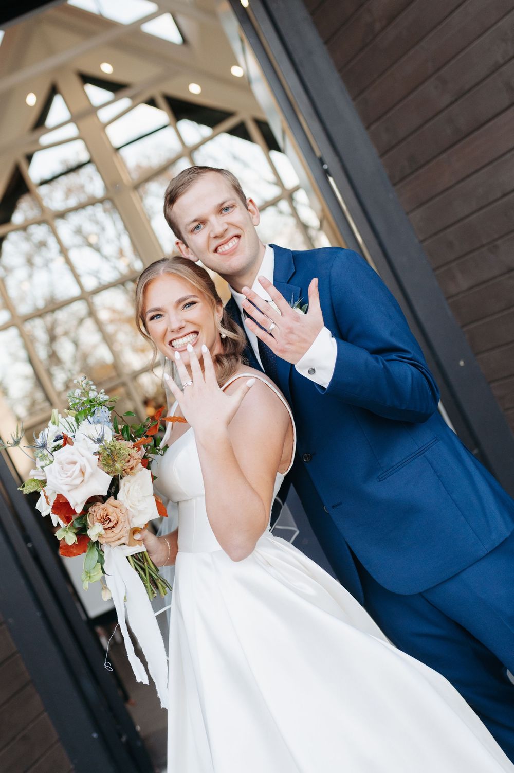 Smiling bride and groom in blue suit show off wedding rings at elegant venue with glass greenhouse backdrop.
