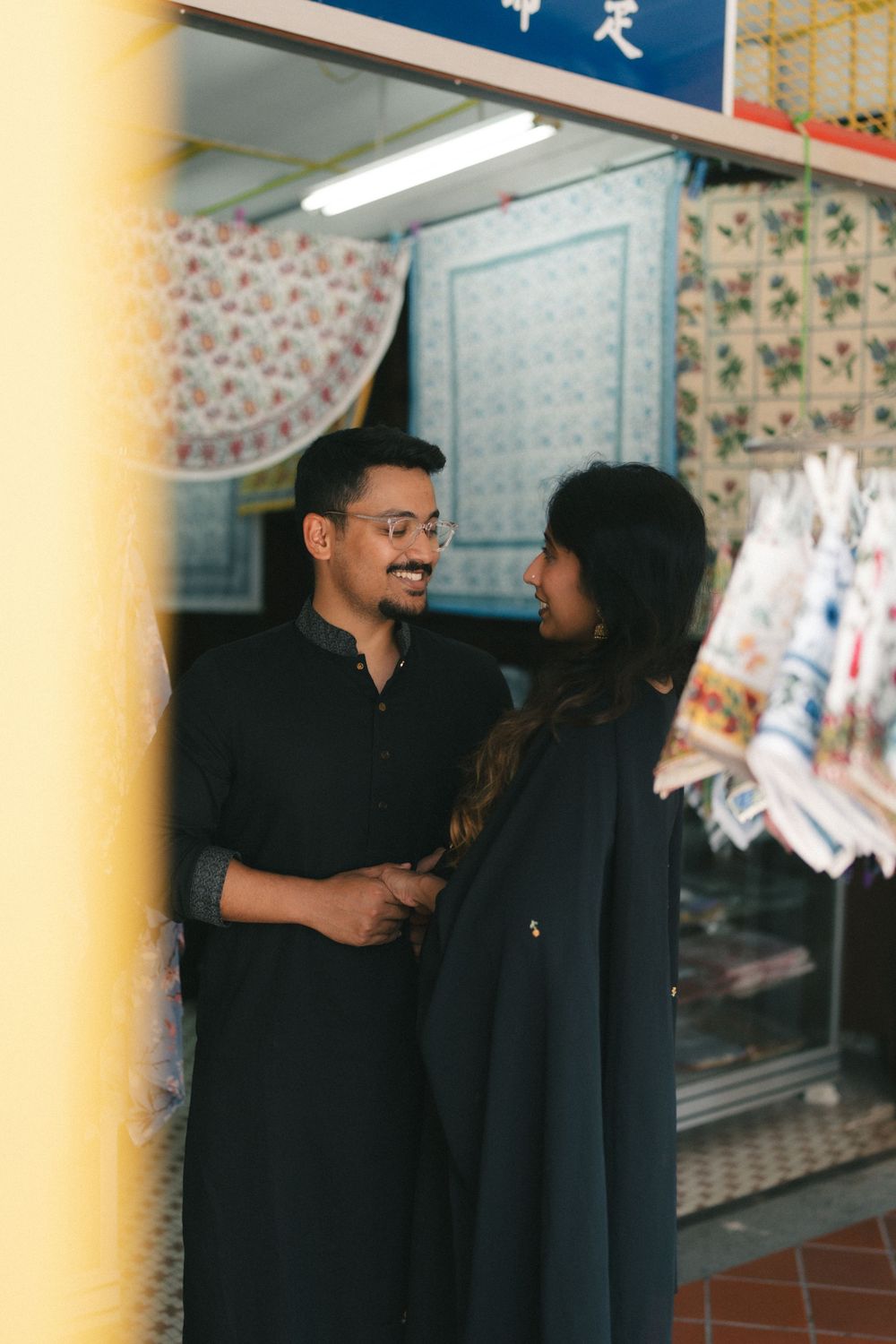 Couple in black traditional attire smiling at each other outside a fabric shop with Chinese signage.