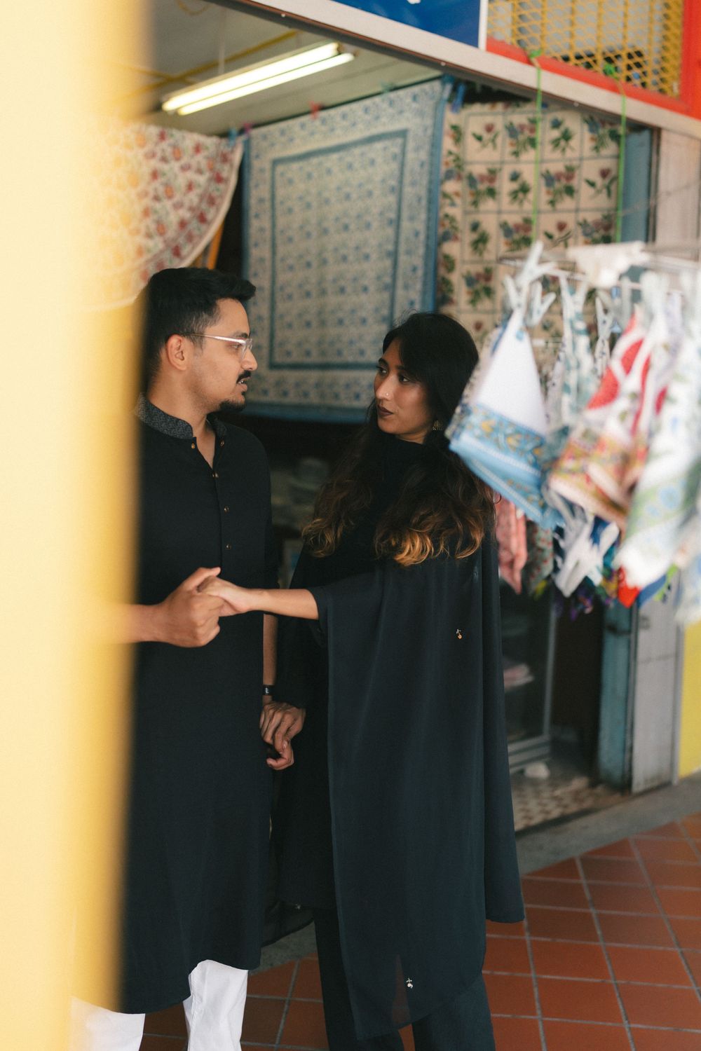 Couple in black traditional attire conversing outside a fabric shop with colorful textiles on display.