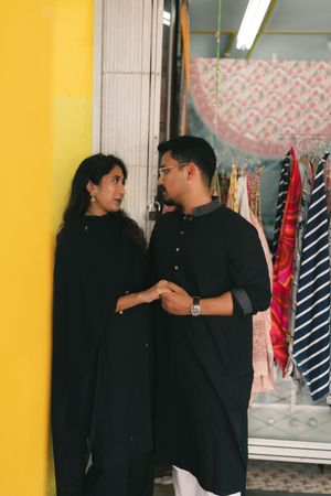 Couple in matching black kurta outfits standing together near a yellow wall outside a clothing boutique.